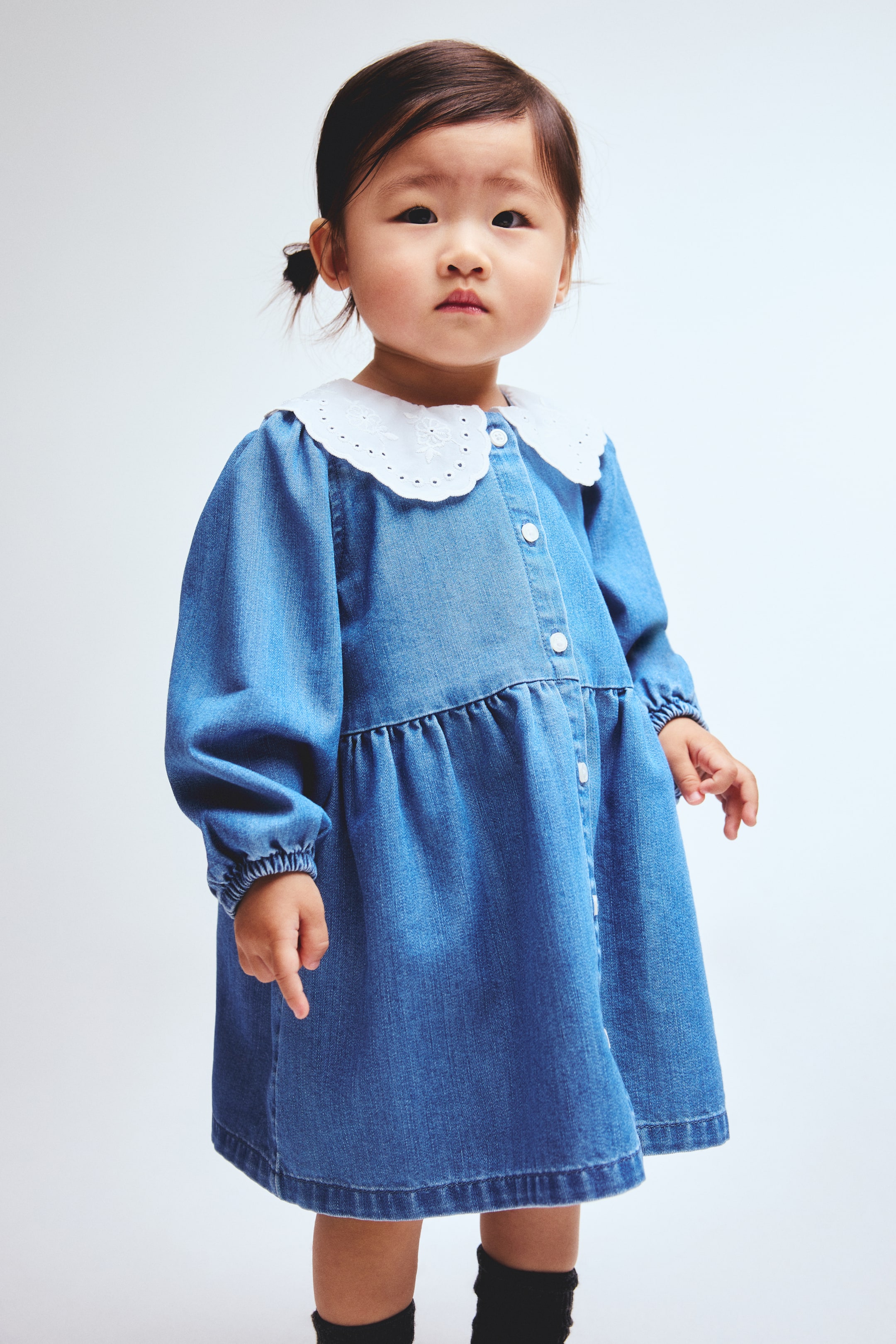 Ampliar la imagen: Un vestido de mezclilla azul para un niño se muestra desde un ángulo frontal. Cuenta con un cuello Peter Pan blanco bordado con un borde festoneado, mangas largas con puños elásticos y un corpiño con botones al frente que se convierte en una falda fruncida con cintura baja.