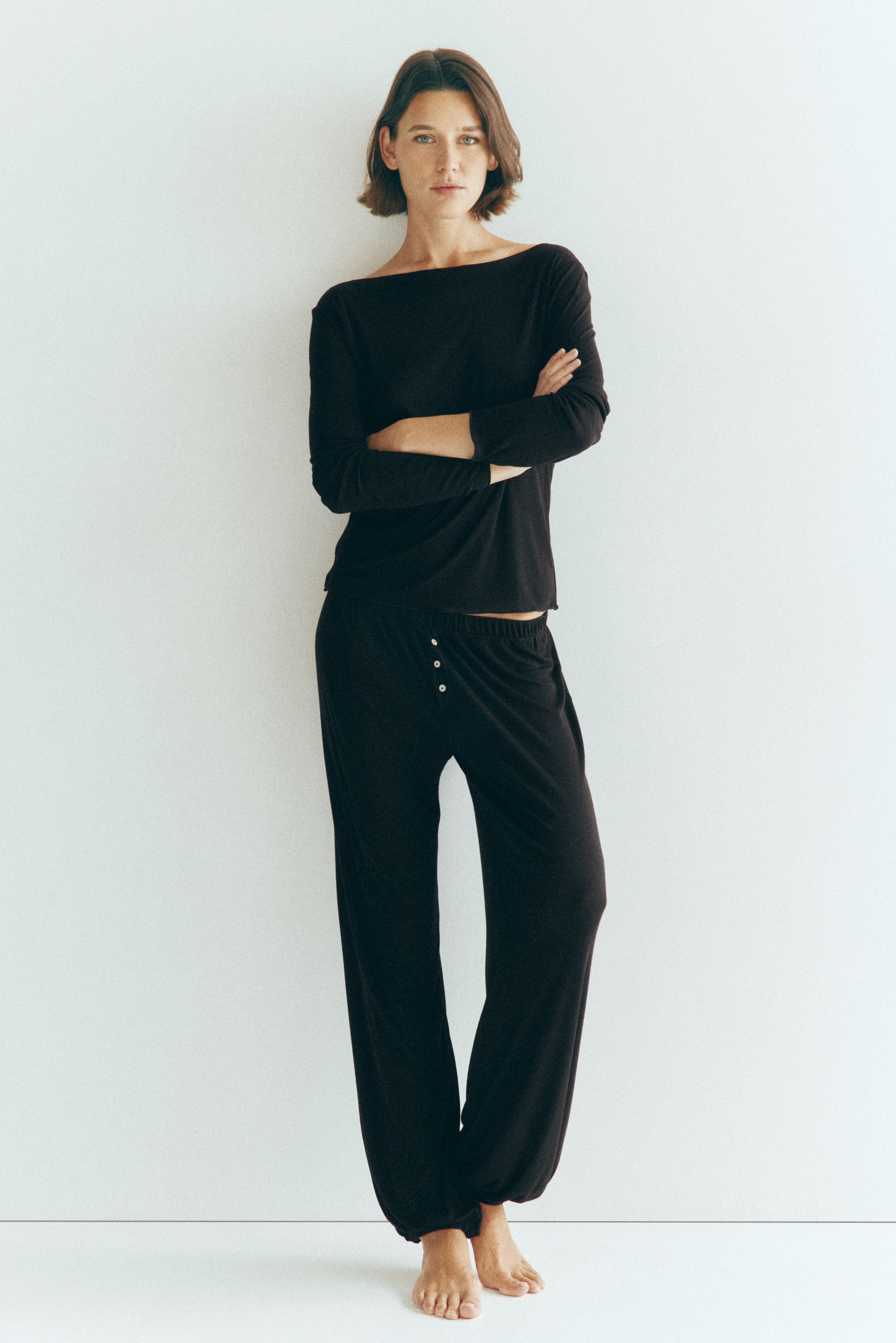 View larger image: The model is wearing a black jersey pyjama set, standing with arms crossed. The long-sleeved top features a boat neck, and the matching trousers feature an elasticated waist, a mock fly with decorative buttons, and balloon legs with elasticated hems.