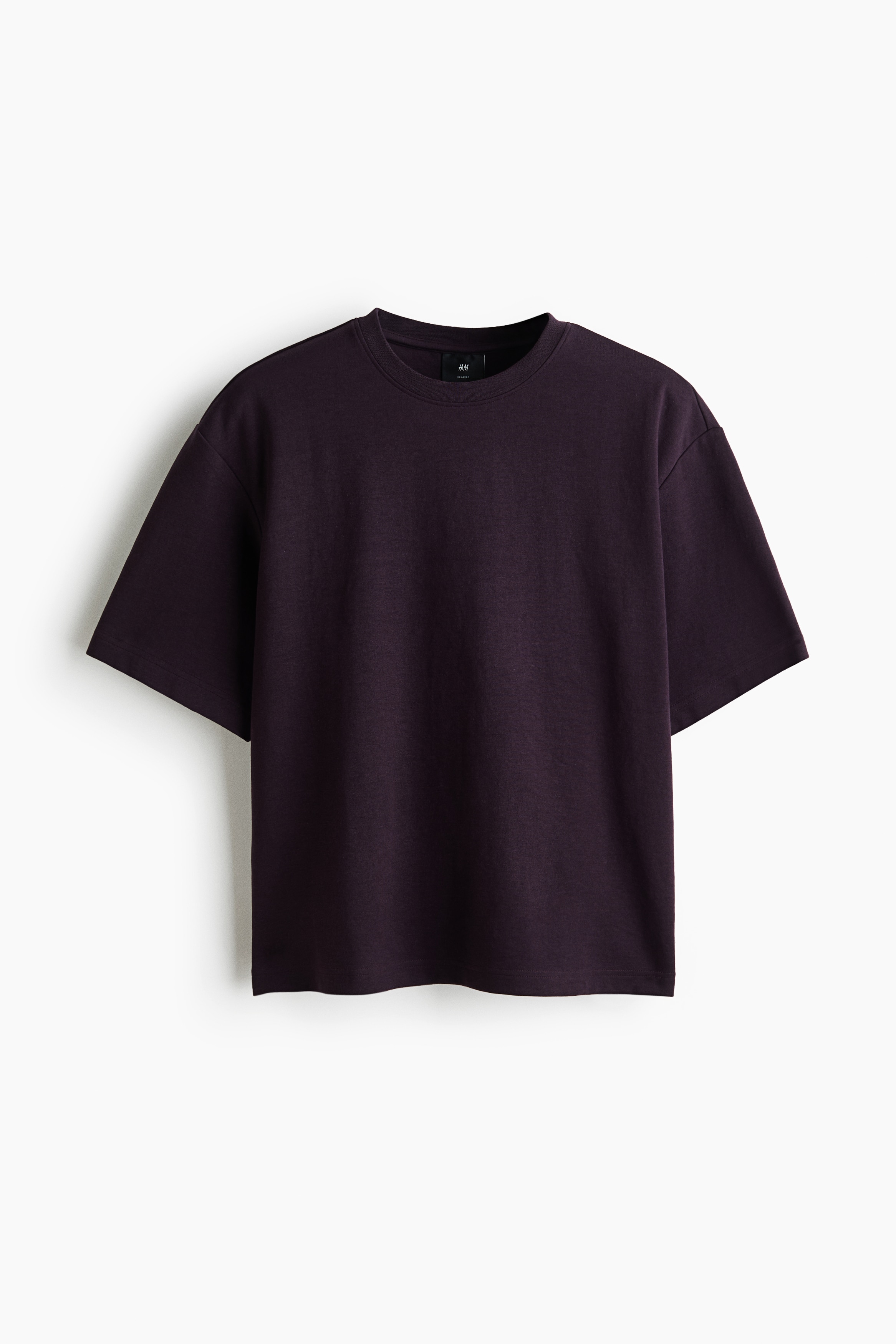 A dark purple heavyweight jersey T-shirt is being worn by the model, featuring a relaxed fit, a round rib-trimmed neckline, dropped shoulders, and a straight hem. The T-shirt is paired with dark grey jeans.
