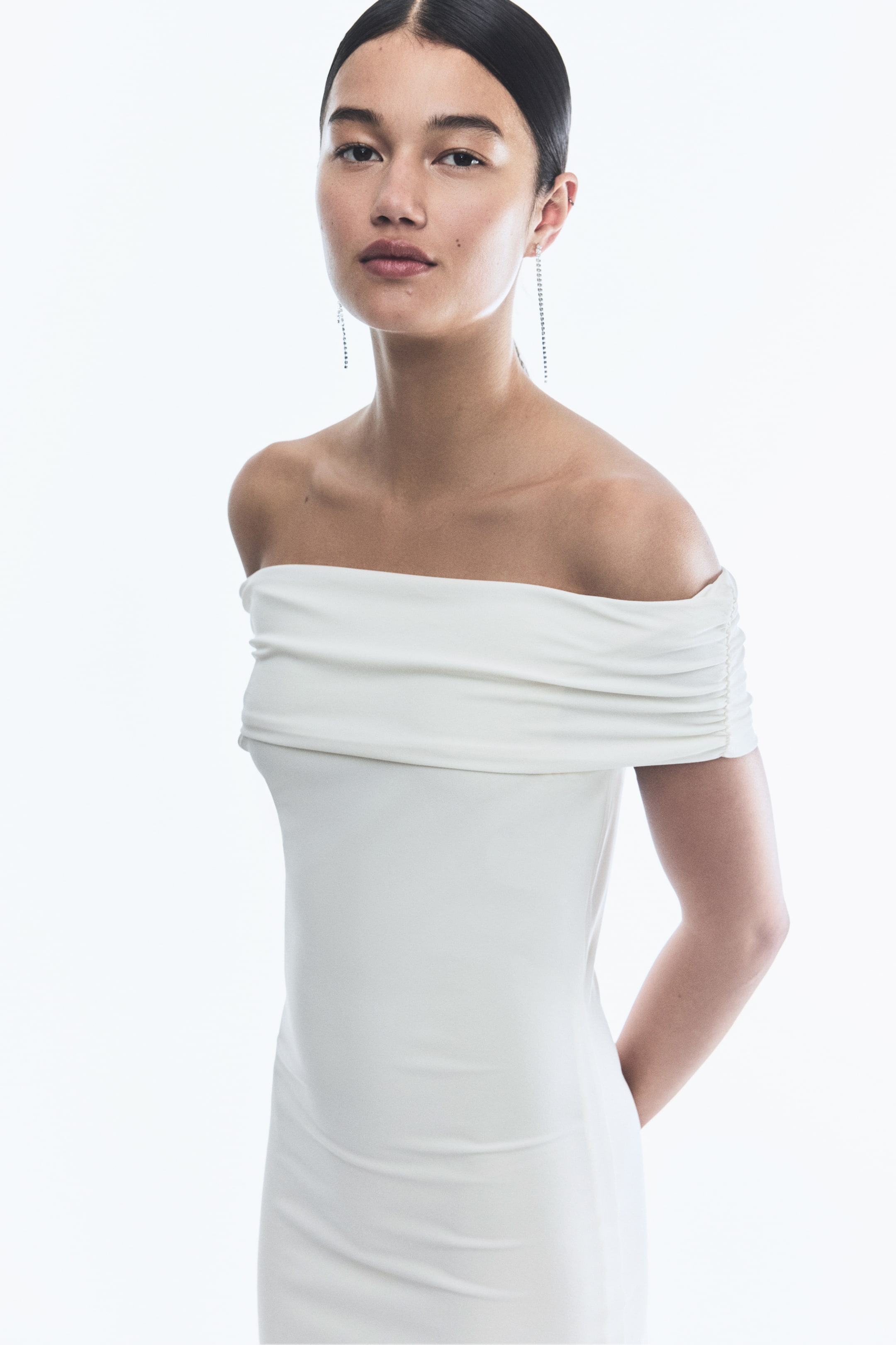 View larger image: A white off-the-shoulder dress is being shown from a slightly front-side angle. It is featuring a draped neckline that is folding over the shoulders, and its short sleeves are gathered with ruching. The dress is closely fitting the body.