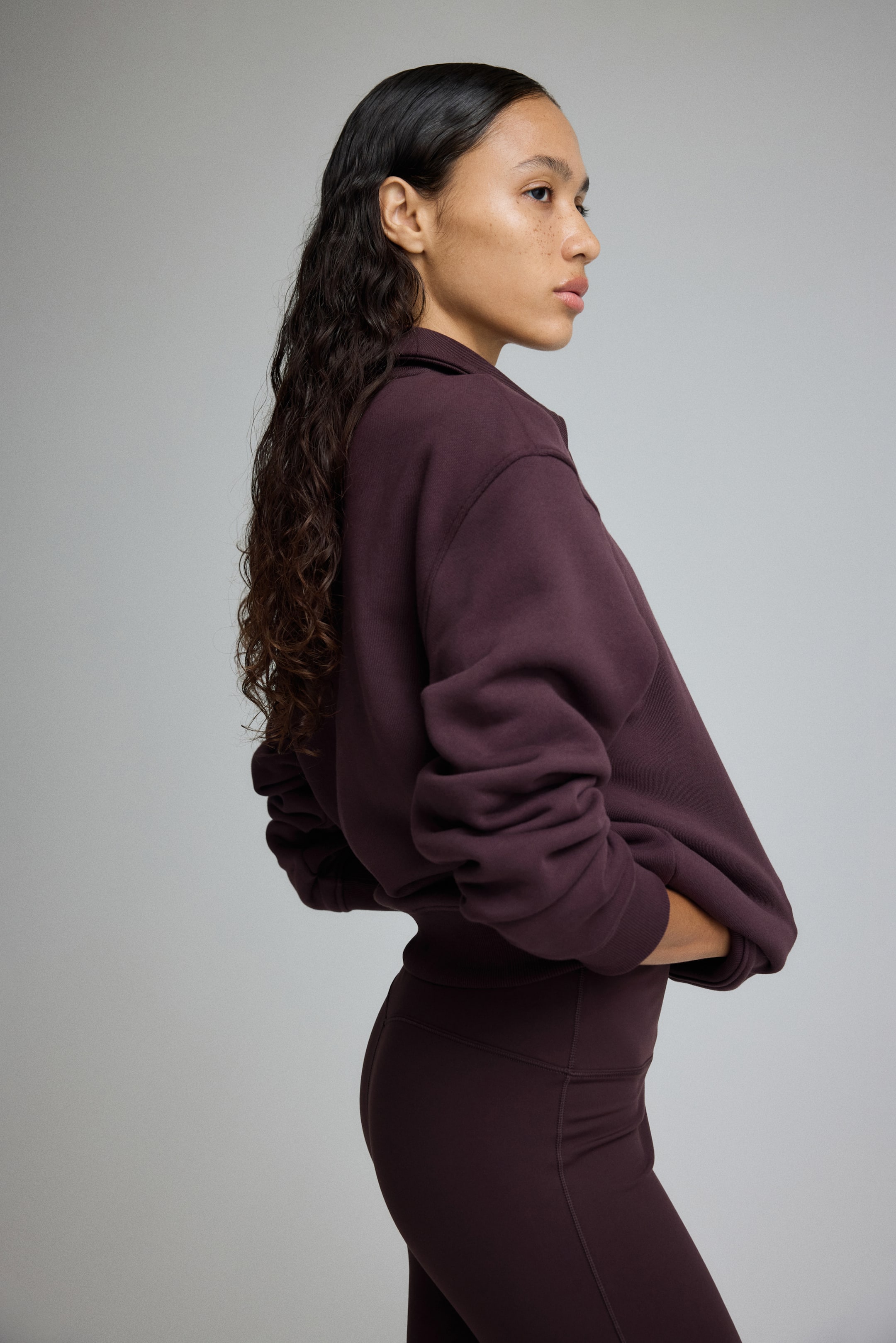 Grotere afbeelding bekijken: Een donker bordeauxrode sweatshirt en bijpassende hoge taille-leggings worden gepresenteerd vanuit een zijprofiel. De sweatshirt heeft een open kraag, volumineuze lange mouwen die bij de manchetten toeknijpen, en een geribbelde zoom die bijeenkomt aan de taille, en de leggings tonen zichtbare naadlijnen langs de heupen.
