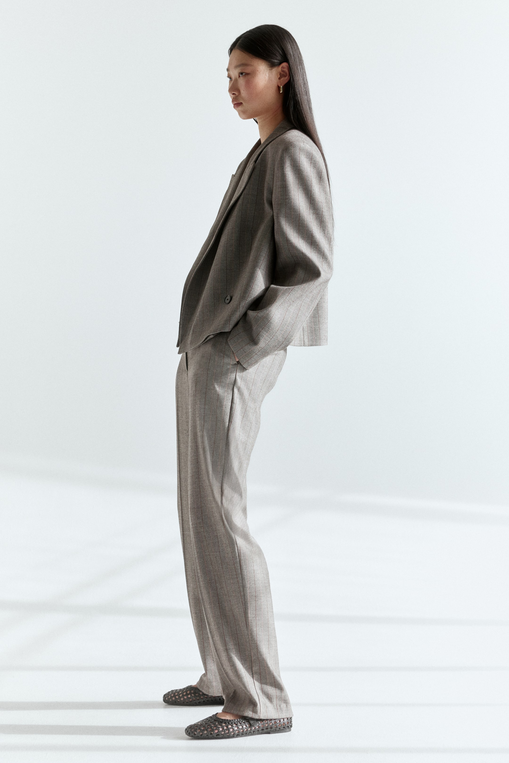 Grotere afbeelding bekijken: Een taupe dubbelrij blazer met krijtstreep en revers met inkeping, gecombineerd met bijpassende wijde broek, wordt vanuit een zijhoek getoond, waarbij de ontspannen pasvorm en drapering worden benadrukt. Donker geweven platte schoenen worden gedragen bij de broek.