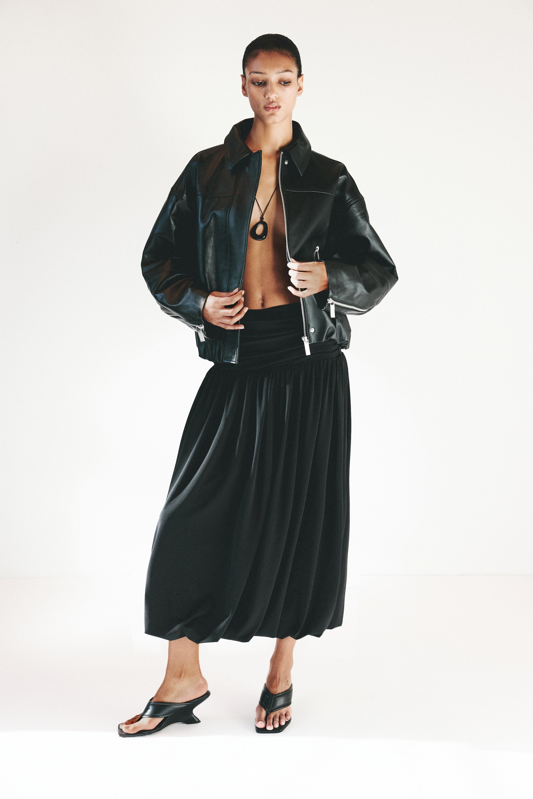 拡大画像を表示: オーバーサイズのフィット感、ポイントカラー、ジッパー付きフロントが特徴のブラックレザージャケットが紹介されています。また、ジッパー付きカフスと伸縮性のある裾が特徴で、リラックスした構造を見せるために開いた状態で保持されています。