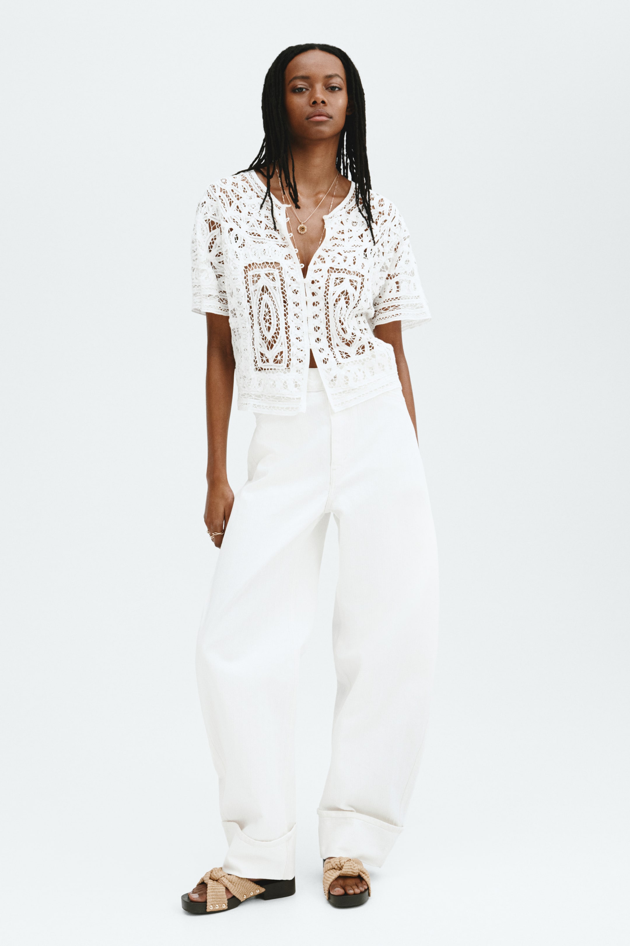 View larger image: A white short-sleeved lace top is displayed front-facing, showcasing detailed openwork designs, a button-front closure, and a cropped hemline. Its sheer material subtly reveals the skin beneath.