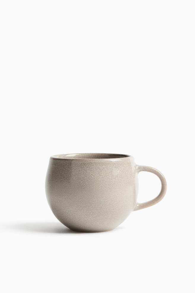 A mug is showcasing its rounded, bulbous body, presented from a slight front angle. It is featuring a light beige speckled glaze and a C-shaped handle on its right side. - 1