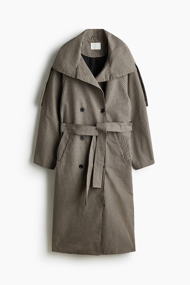 An oversized, double-breasted trench coat in a brown-and-black gingham pattern is shown facing front, displaying a wide collar, raglan sleeves with shoulder tabs, and a matching wide tie belt at the waist. The coat is featuring dark double-breasted buttons, welt front pockets, and a black lining visible at the open neckline. - 1