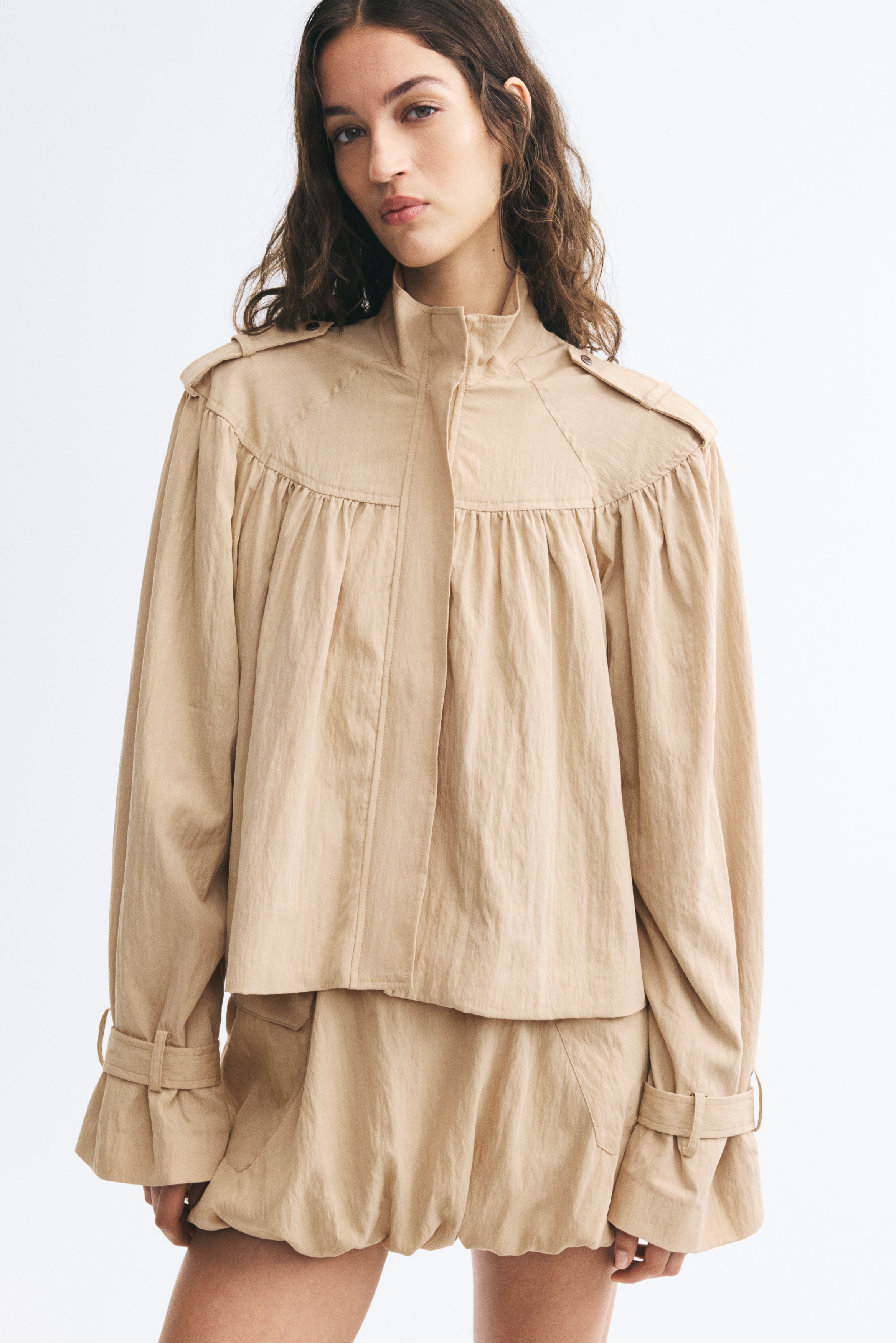 A beige cropped jacket is hanging front-facing, featuring a high stand-up collar, shoulder epaulets, and a concealed front placket. The design showcases prominent gathering below the yoke that creates a voluminous drape throughout the body and wide sleeves, finished with adjustable cuff straps.