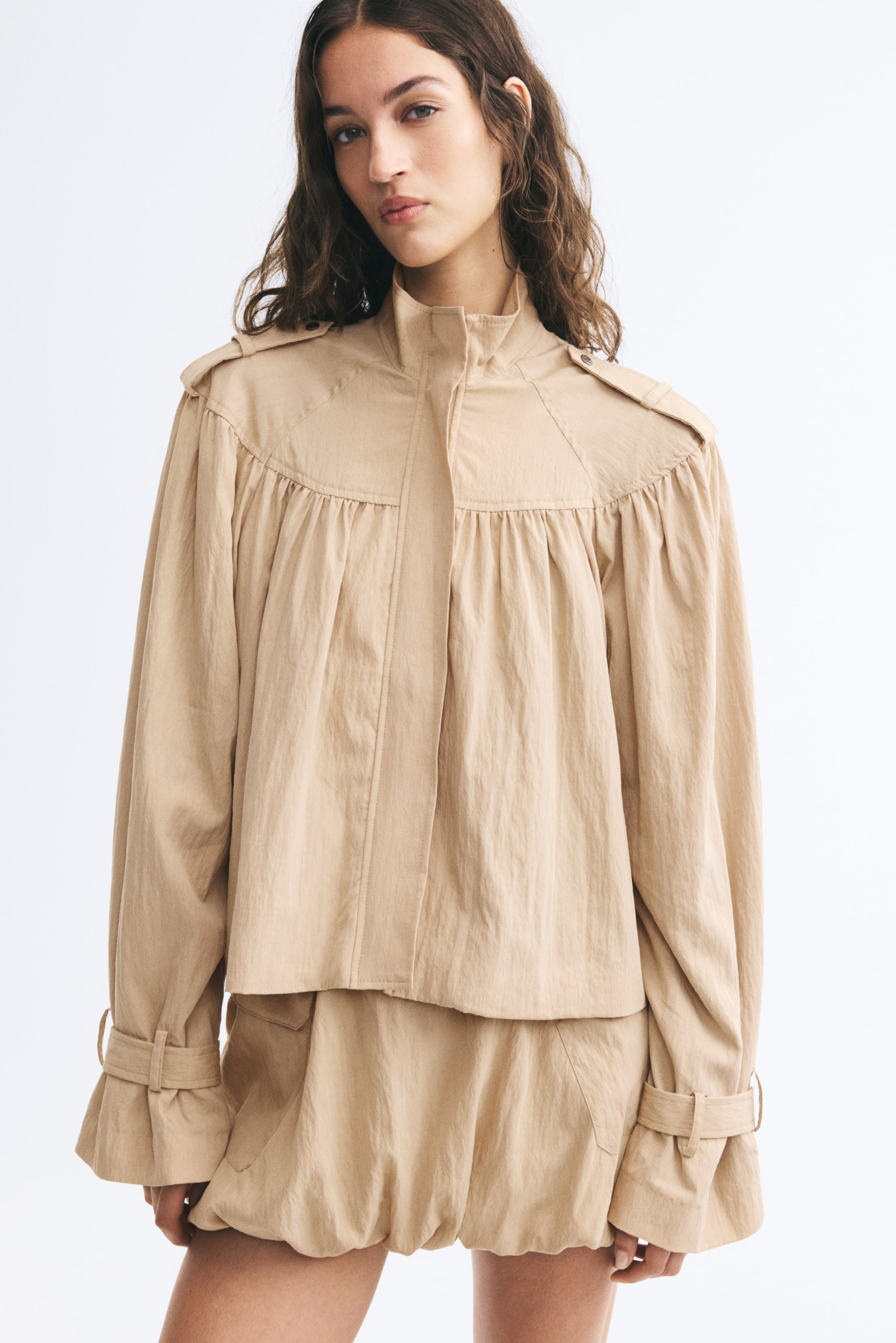 View larger image: A short beige jacket in a textured, crinkled weave is worn by the model, featuring a high funnel collar, gathered front yoke, and shoulder tabs. The jacket is styled with long raglan sleeves with adjustable cuff tabs and paired with matching beige shorts.