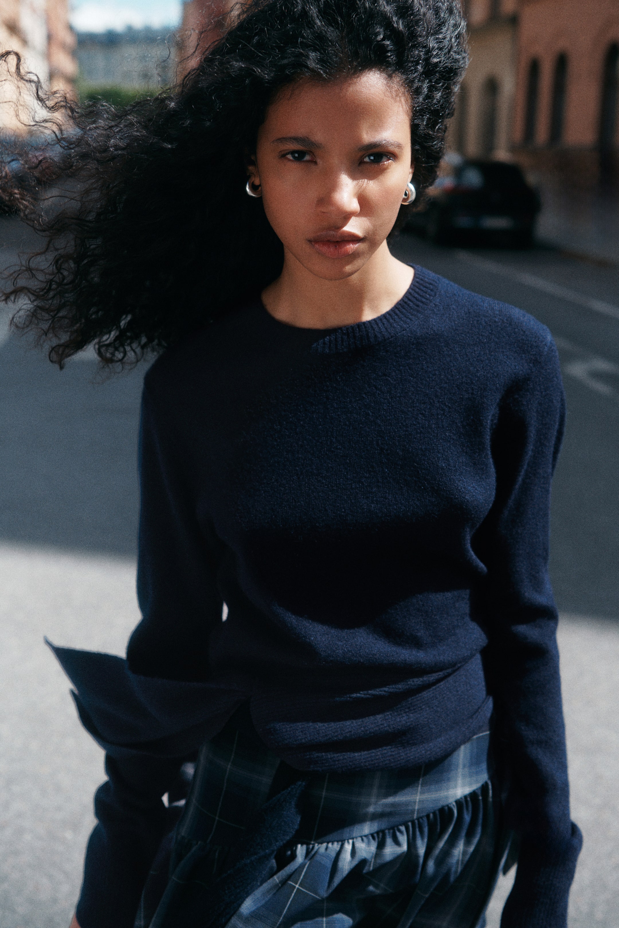 View larger image: A dark navy blue knit sweater with a ribbed crew neckline and a wide tie detail at the front hem is being worn by the model. The sweater is paired with a blue plaid skirt.