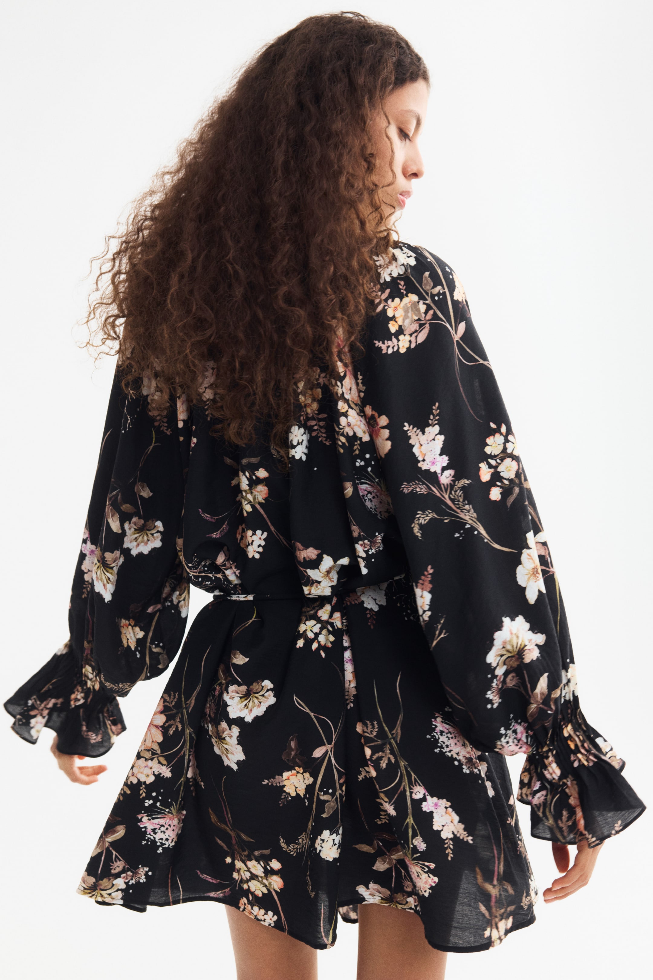 Größeres Bild ansehen: Ein kurzes Kleid in lockerer Passform aus schwarzem Stoff mit Blumenprint wird vom Model getragen und ist in einer dreiviertel Rückansicht zu sehen. Das Kleid verfügt über lange, voluminöse Raglanärmel mit gerafften Handgelenken und angesetzten Volants an den Ärmelabschlüssen sowie eine geraffte Taille.