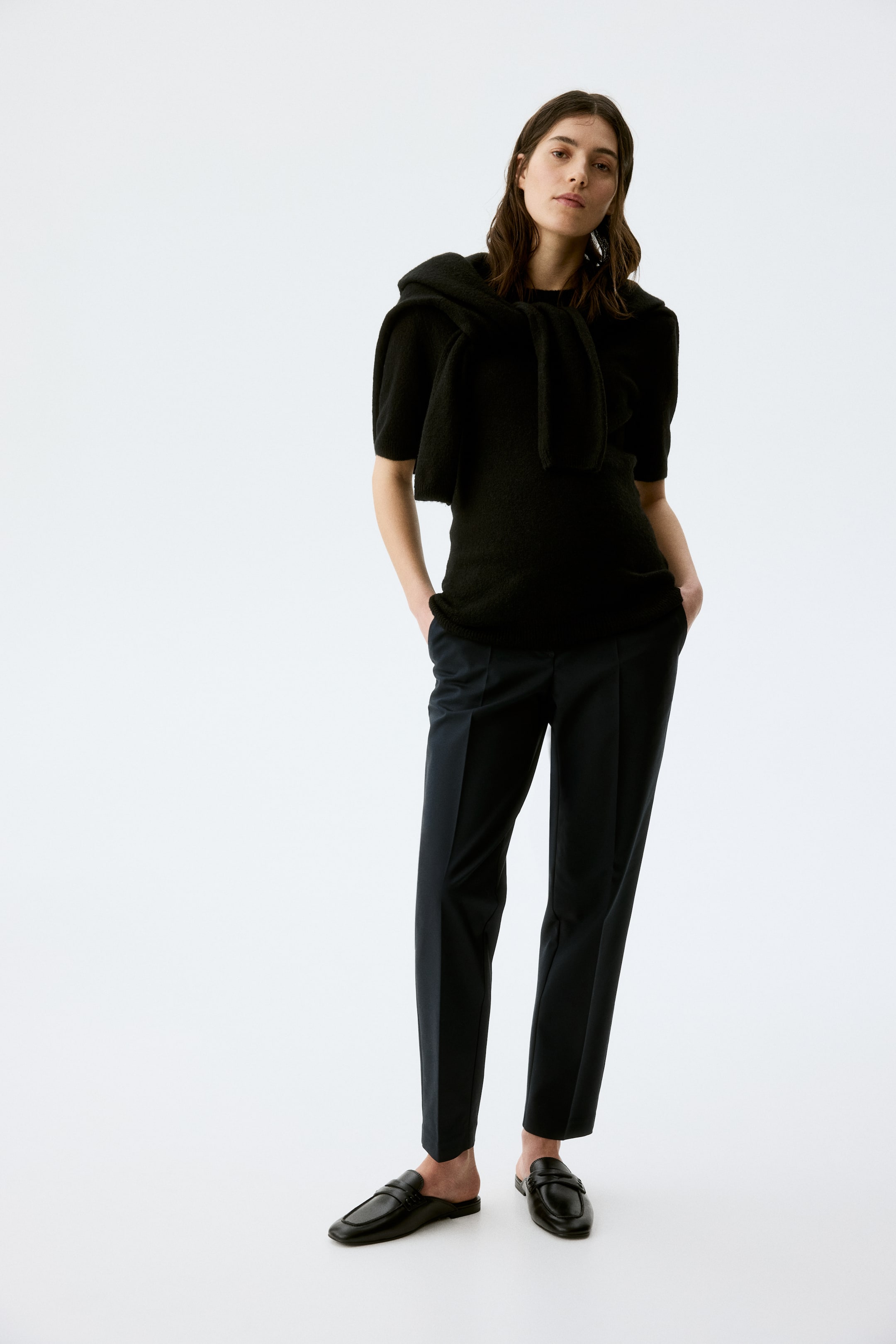 Προβολή μεγαλύτερης εικόνας: Ένα μαύρο κοντομάνικο πλεκτό τοπ παρουσιάζεται, συνδυασμένο με ένα μαύρο πλεκτό πουλόβερ δεμένο στους ώμους. Μαύροι ίσιοι παντελόνια με μπροστινή πιέτα και μήκος μέχρι τον αστράγαλο, και μαύρα δερμάτινα μουλ λόφερς με τετράγωνη μύτη και μεταλλικές λεπτομέρειες αγκράφας φοριούνται.