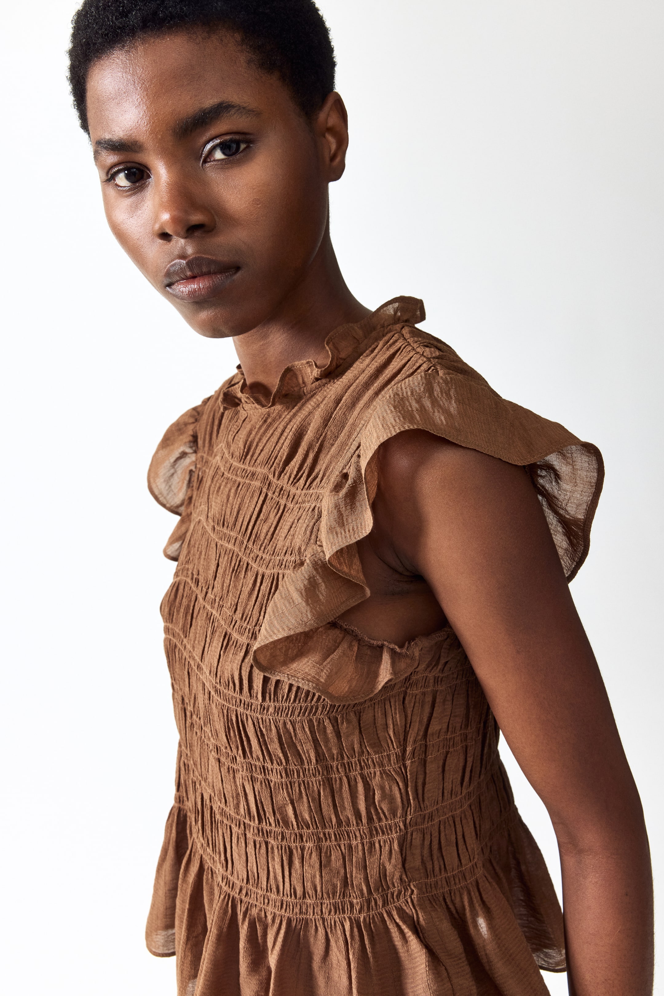 Ingrandisci l'immagine: La modella indossa un top peplo arricciato in marrone. Presenta un collo alto con volant, maniche a cappuccio con volant e un ampio arricciamento lungo tutto il corpetto, che si apre in un peplo.