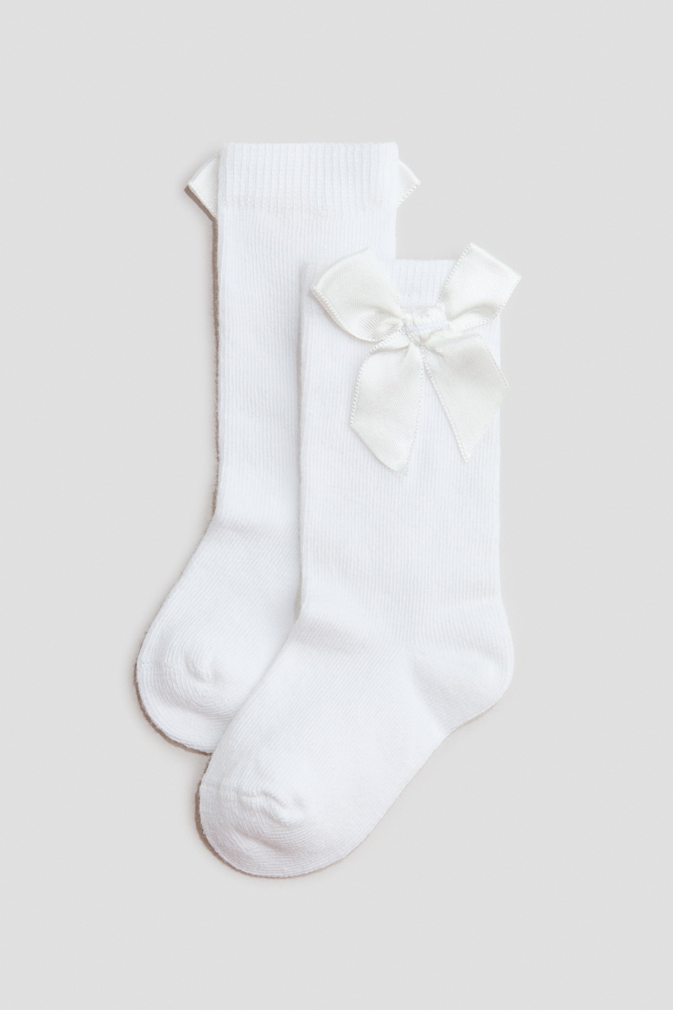 Agrandir l'image: Deux chaussettes blanches en laine présentent de face, fabriquées à partir d'un tissu tricoté fin et doux. Chaque chaussette est dotée d'une partie supérieure côtelée et élastique, et un nœud en satin blanc est positionné juste en dessous du rebord.