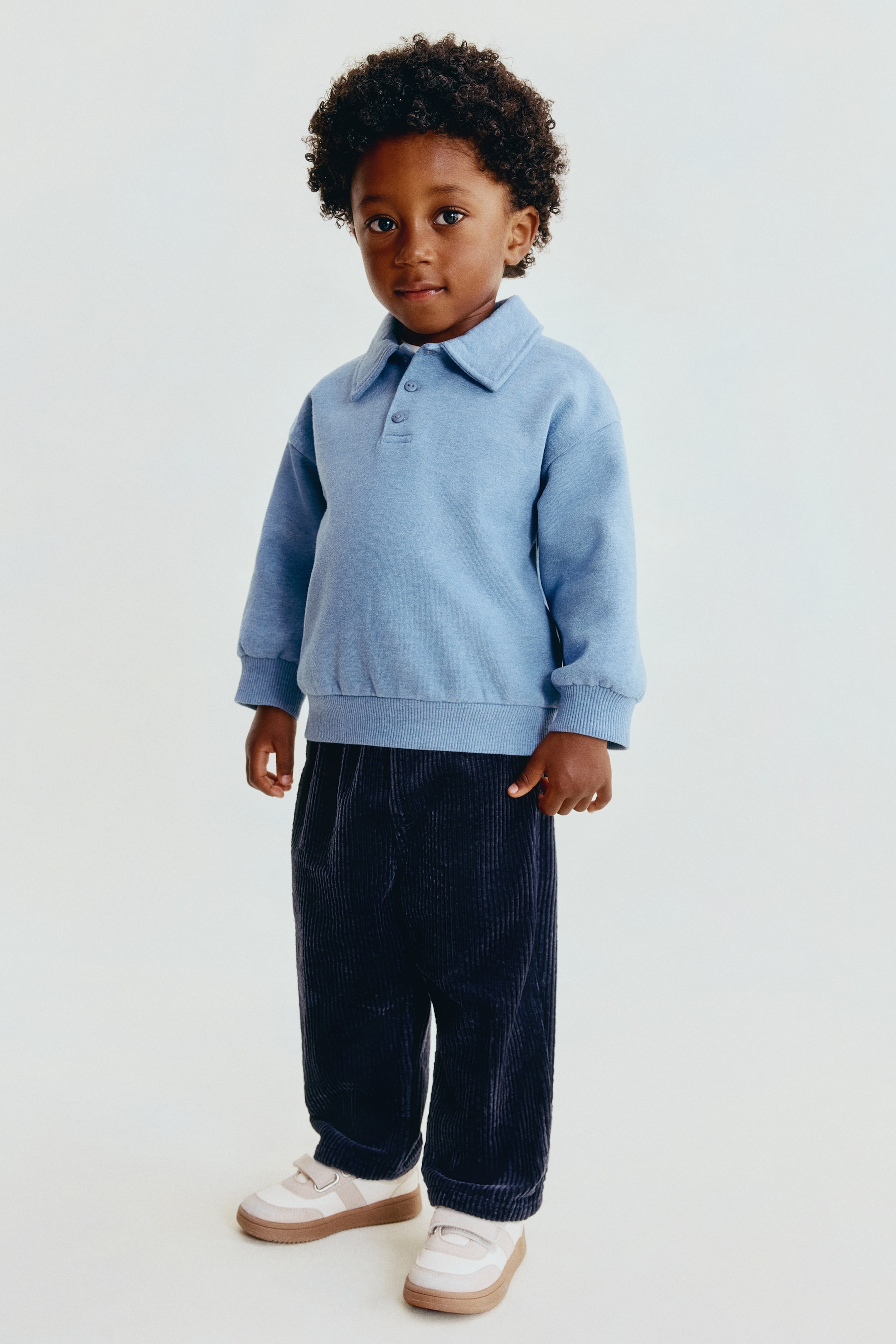 Visualizza l'immagine ingrandita: Un bambino indossa pantaloni in velluto a coste blu navy scuro, ripreso frontalmente, mettendo in risalto la vestibilità ampia e dritta e la texture verticale a coste. I pantaloni sono portati in vita con una fascia elastica arricciata, parzialmente coperta da una felpa azzurra con colletto.