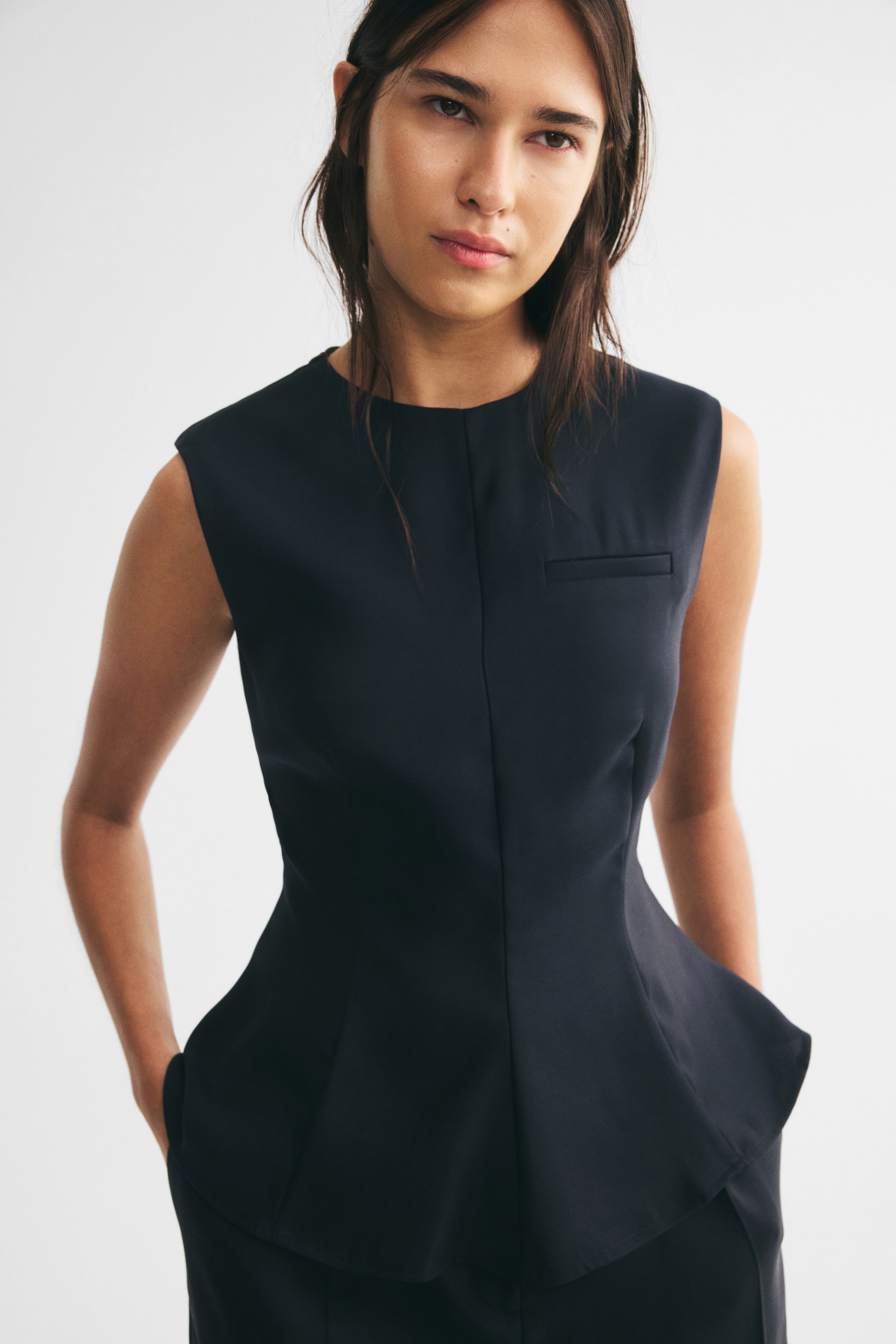 View larger image: A dark navy sleeveless peplum top is worn by the model, featuring a round neckline, a prominent vertical seam down the center front, a mock welt pocket on the chest, and a flared waist. The model is standing with her hands in the pockets of matching dark trousers.