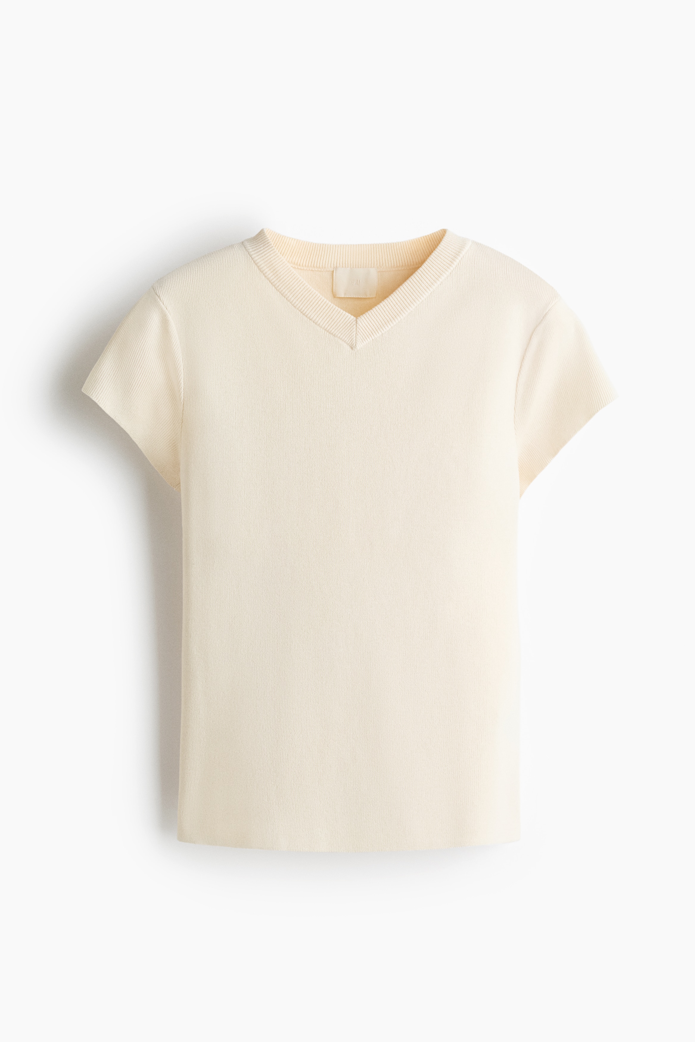 A cream-colored, fitted ribbed-knit tee is being worn by the model in a side-profile view. The short-sleeved top features a fine ribbed texture, a round neckline, and a snug fit.