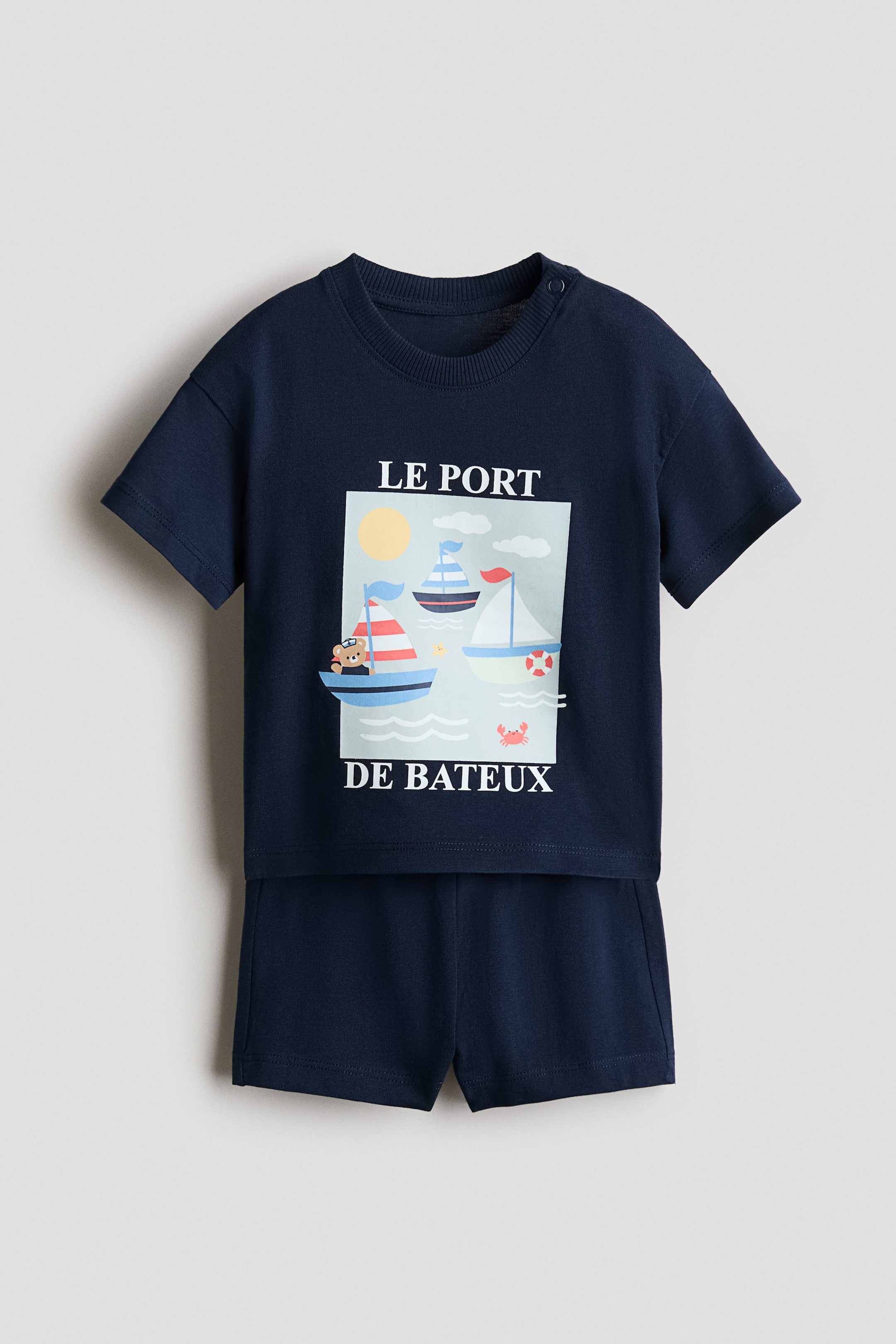 拡大画像を表示: ネイビーブルーのコットン製2点セットで、半袖Tシャツと同色のショーツが揃い、商品は正面から提示されています。Tシャツはクルーネック仕様で右肩にスナップボタンが2つ付き、中央には3隻の船、クマ、太陽、雲、カニを描いた航海モチーフのグラフィックと「LE PORT DE BATEAUX."」という文字が配されています。