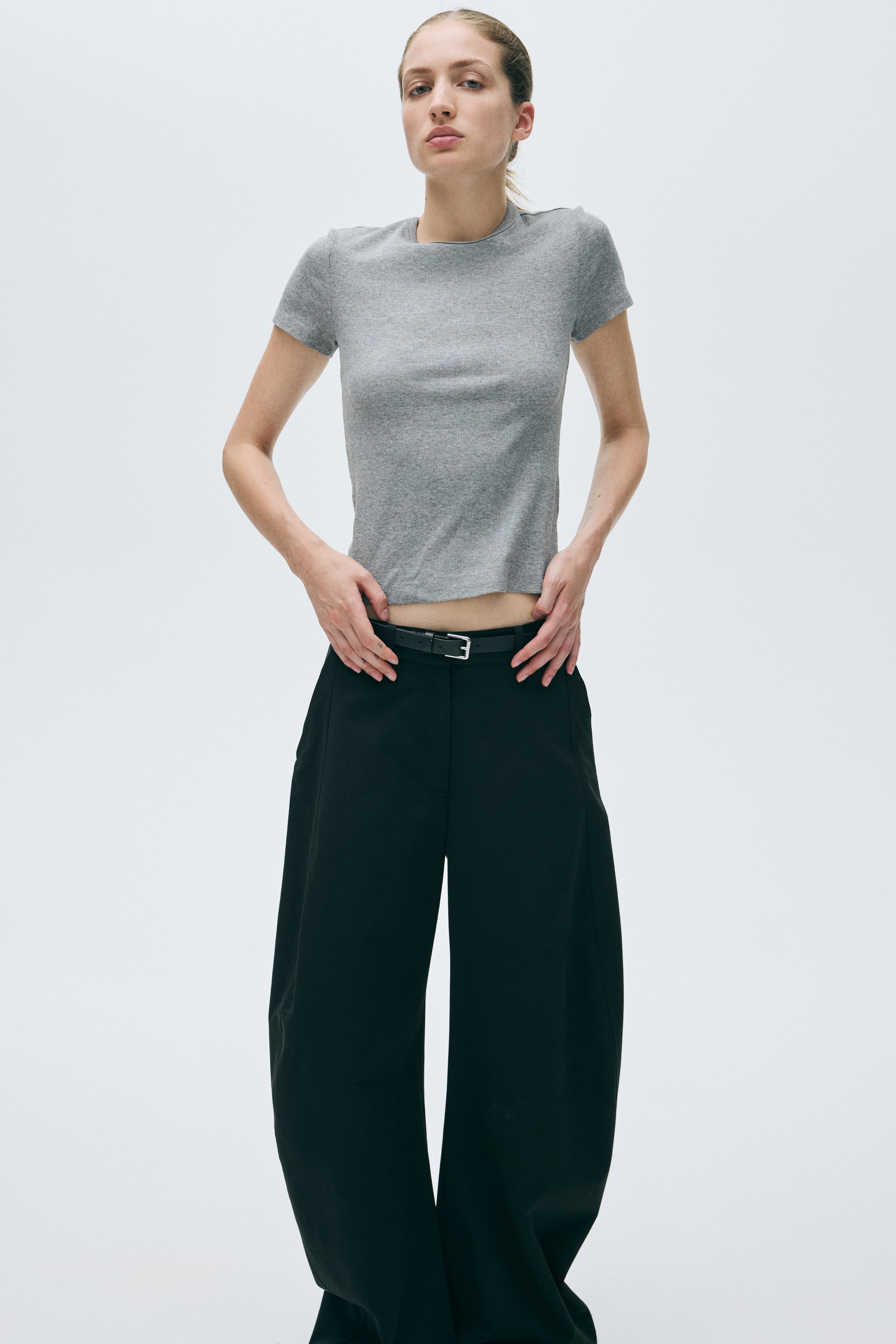 View larger image: A fitted grey cropped crew neck t-shirt with short sleeves is showcased from a front angle, paired with black wide-leg trousers that are cinched at the high waist by a thin black belt featuring a silver buckle.