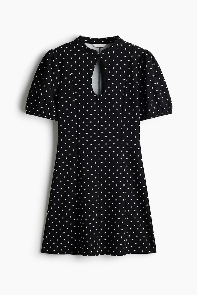 A black A-line dress with a white polka dot pattern is shown from the front, featuring a band collar, a central keyhole cutout at the chest, and short puff sleeves. - 1