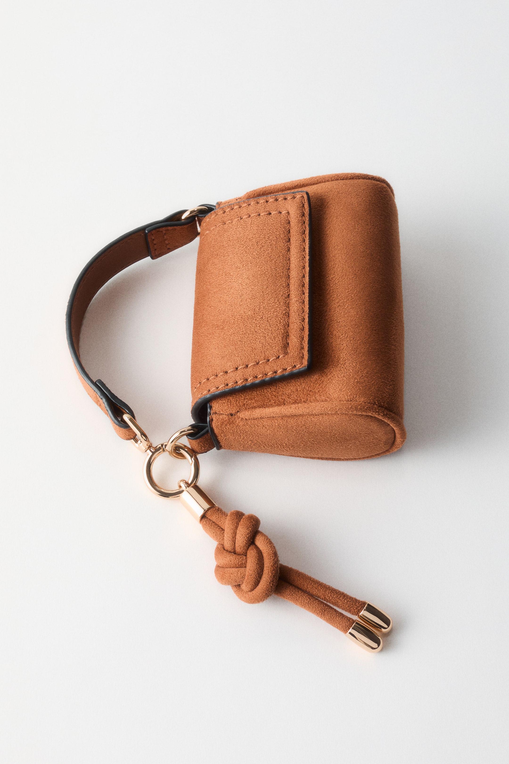 Ingrandisci l'immagine: Una piccola borsa in similpelle scamosciata marrone è inclinata verso destra, caratterizzata da una chiusura a patta e un sottile cinturino marrone scuro. Un hardware dorato collega il cinturino alla borsa, e un decorativo nappina annodata con terminali dorati abbinati pende da essa.