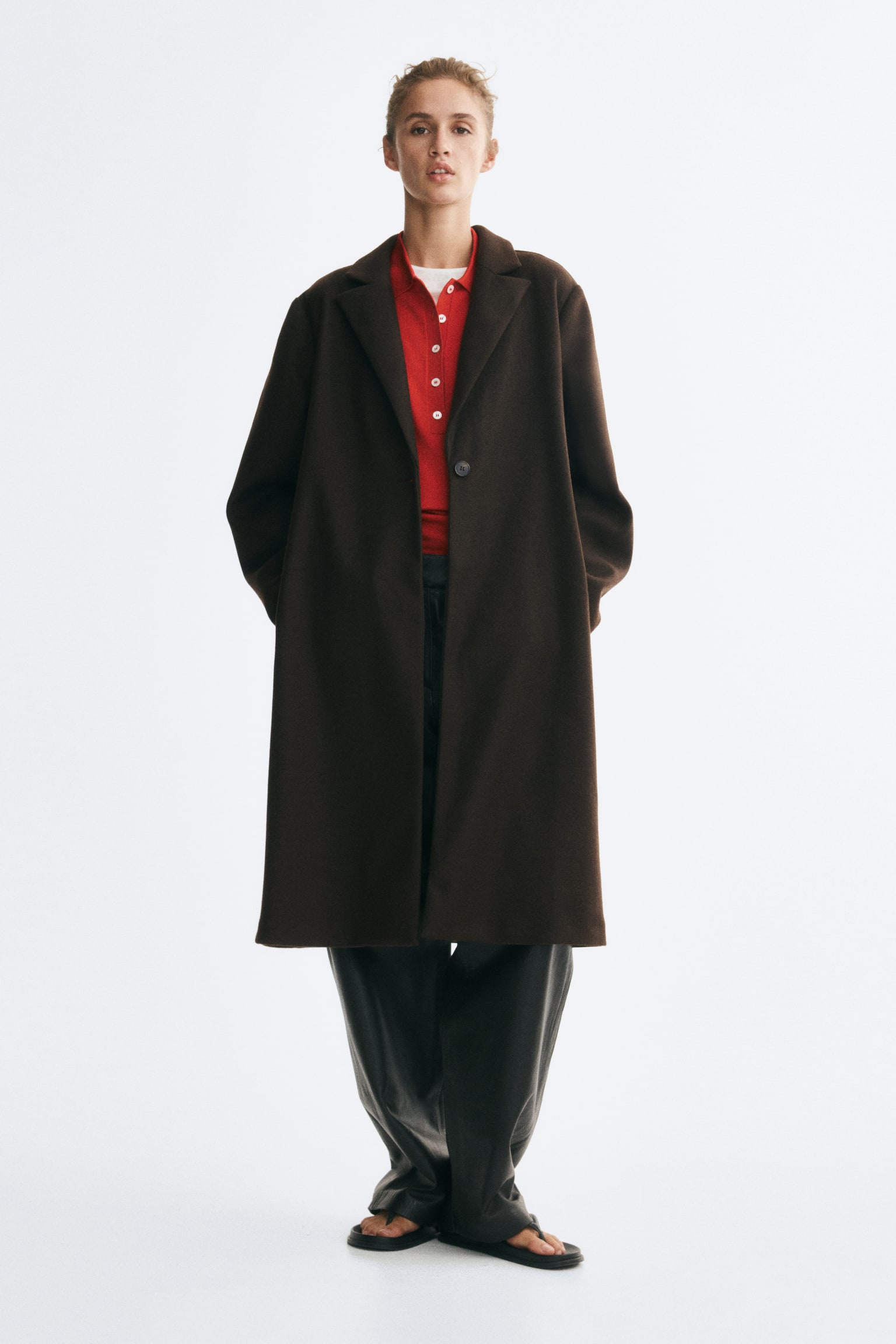 A knee-length, single-breasted coat in dark brown felted fabric is worn by the model over a red polo shirt and dark, wide-leg trousers. The coat features wide notch lapels, a single front button, and side pockets, with the model resting their hands in them. - 1