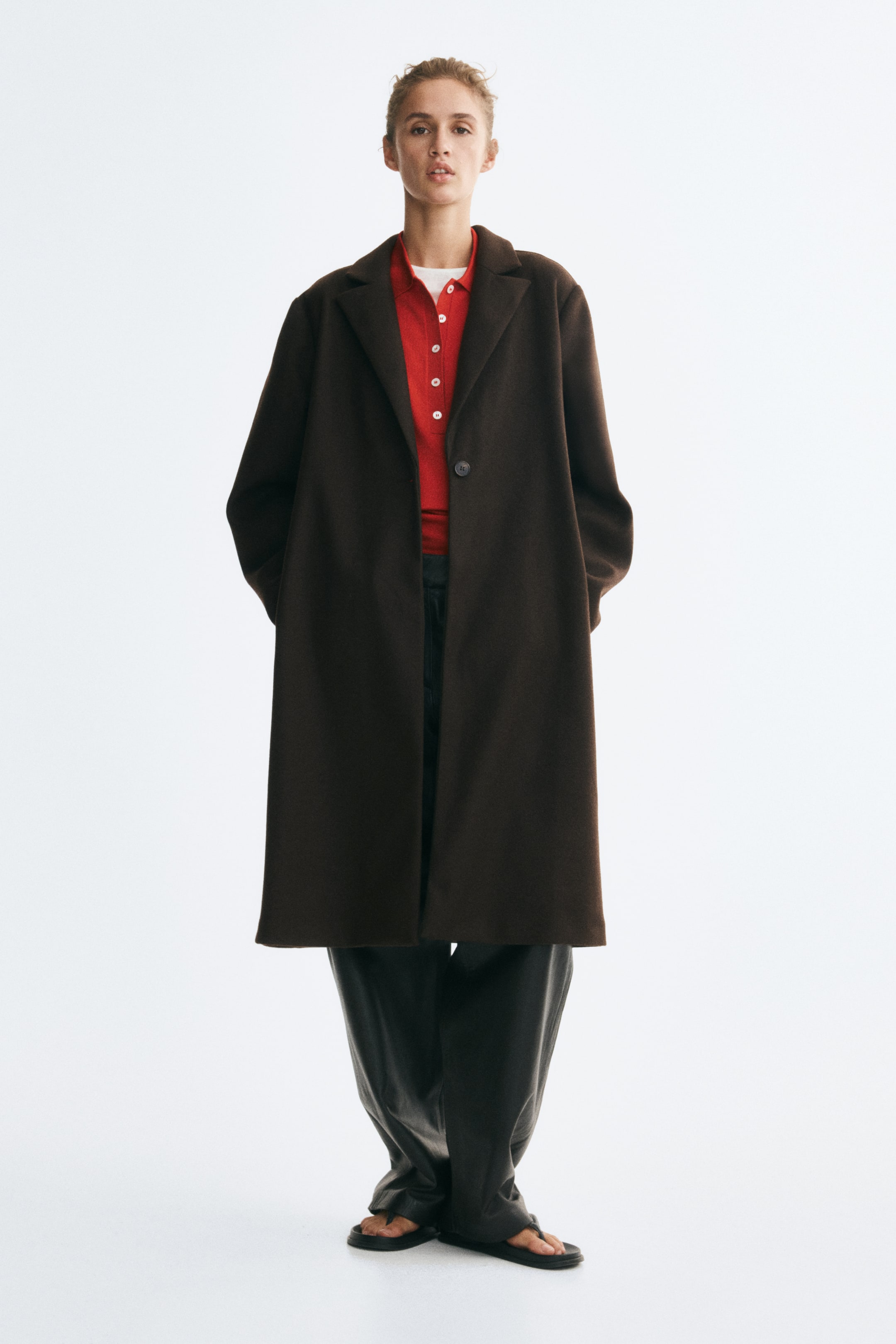 Agrandir l'image: Un manteau mi-long à simple boutonnage en tissu feutré brun foncé est porté par le mannequin par-dessus un polo rouge et un pantalon large foncé. Le manteau présente de larges revers crantés, un seul bouton sur le devant et des poches latérales dans lesquelles le mannequin repose ses mains.