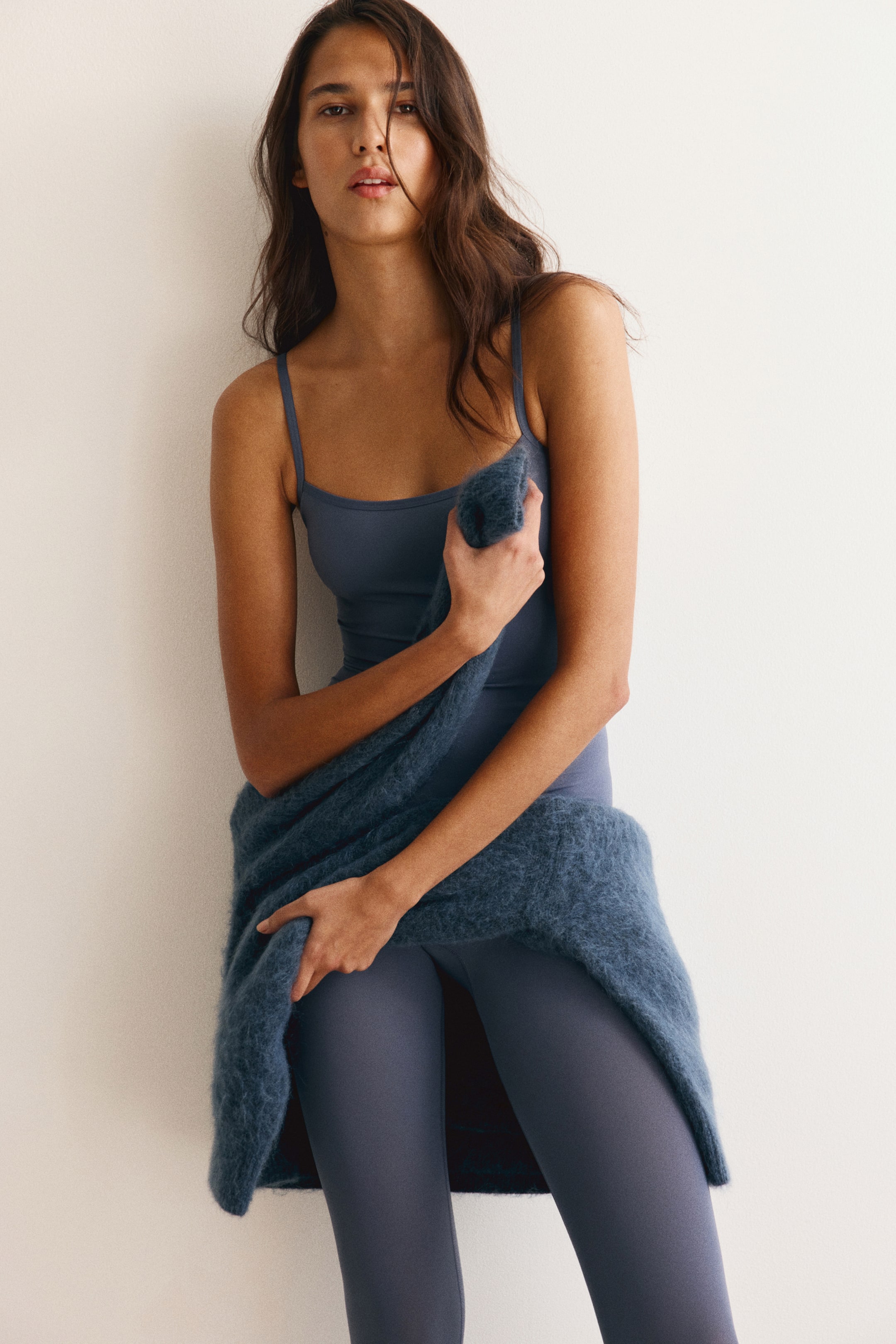 View larger image: A blue stretch jersey unitard with a straight neckline and thin shoulder straps is worn by the model. The model is holding a matching fuzzy knit sweater draped around the waist.