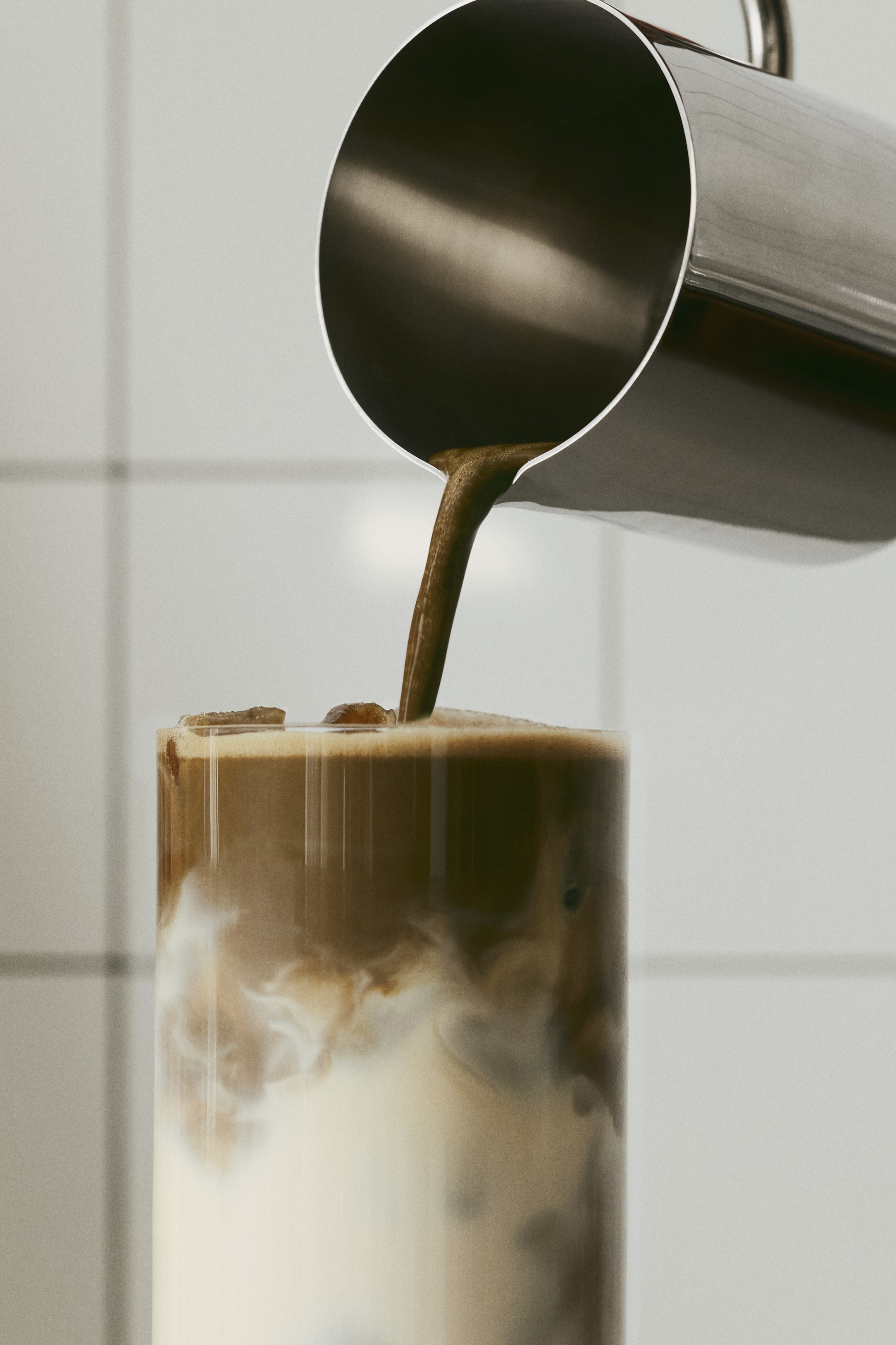Voir une image plus grande: Un petit pichet à lait en acier inoxydable verse un filet de liquide foncé dans un verre transparent. Le pichet est incliné vers le bas, présentant un extérieur lisse et réfléchissant ainsi qu’un bec verseur précis.