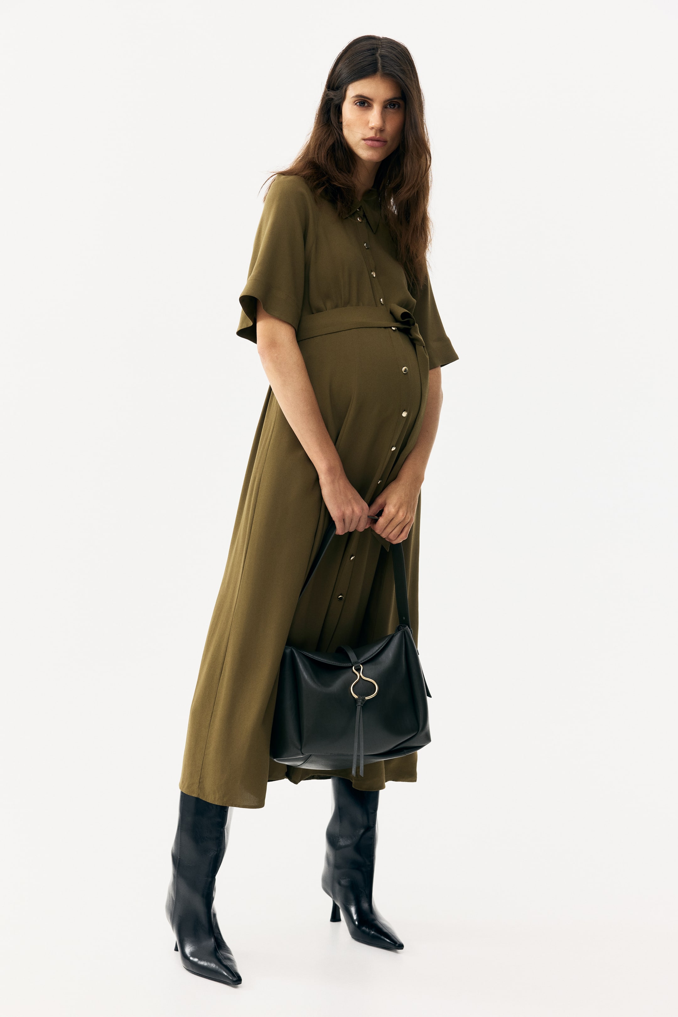 Ver imagem maior: Um vestido-camisa midi verde azeitona exibe uma gola pontiaguda, mangas curtas e largas, e uma frente totalmente abotoada com botões em tom dourado. O vestido ajusta-se na cintura com um cinto de laço a condizer, acomodando uma barriga de grávida visível, e flui até um comprimento midi.