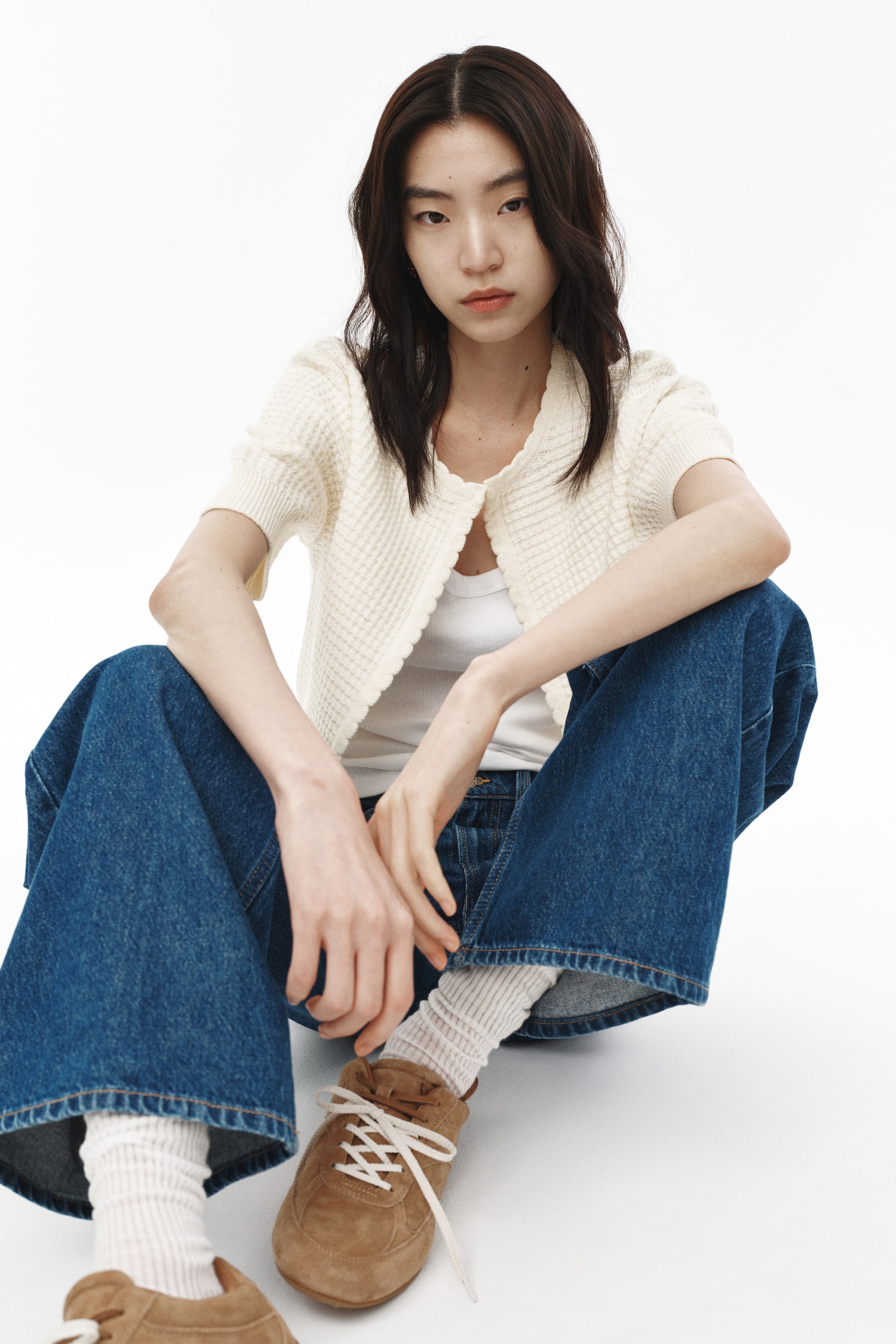 Xem ảnh lớn hơn: Chiếc áo cardigan màu kem tay ngắn được đặt hướng về phía trước, hiển thị họa tiết dệt waffle, cổ tay áo bo gân và có một chiếc nút cài duy nhất ở phía trên, được phối bên ngoài áo tank top màu trắng.