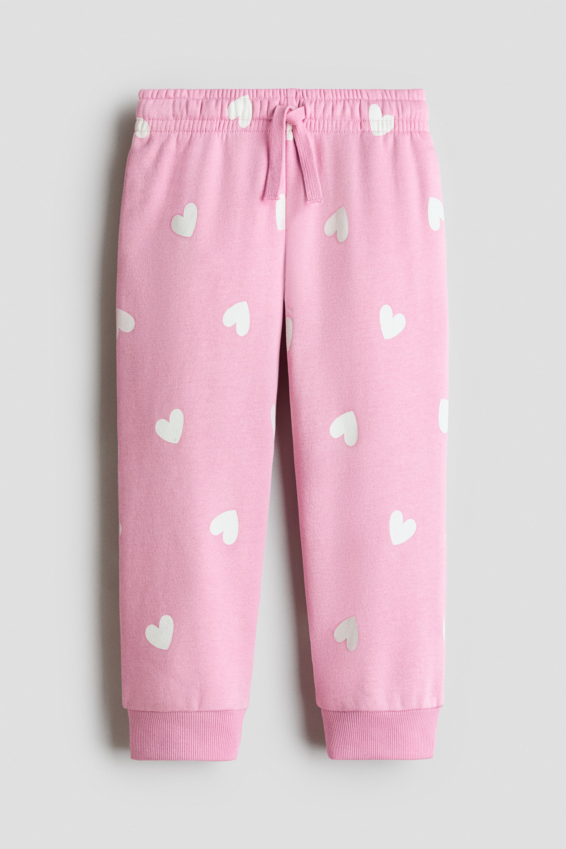 View larger image: Pink sweatpants are laid flat, featuring an all-over pattern of white hearts. They are designed with an elasticized waistband, including a pink drawstring, and ribbed cuffs at the ankles.
