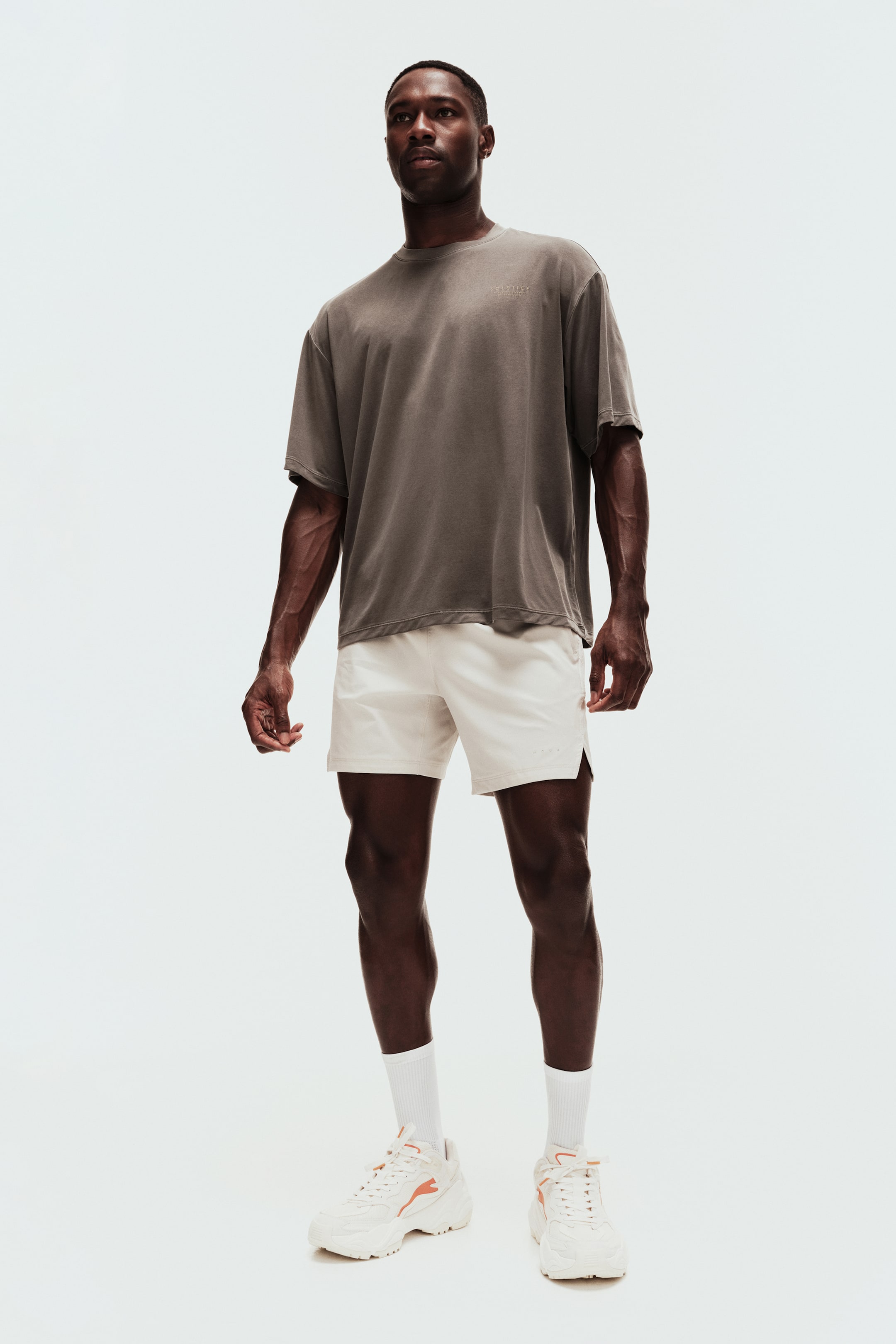 Ver imagem maior: Uma t-shirt oversized cinzento-escuro com um pequeno logótipo bordado está a ser usada, combinada com calções desportivos creme claro que apresentam uma bainha dividida lateral. Meias brancas de cano alto estão a ser usadas com ténis brancos volumosos, acentuados com detalhes laranja, completando o conjunto virado para a frente.