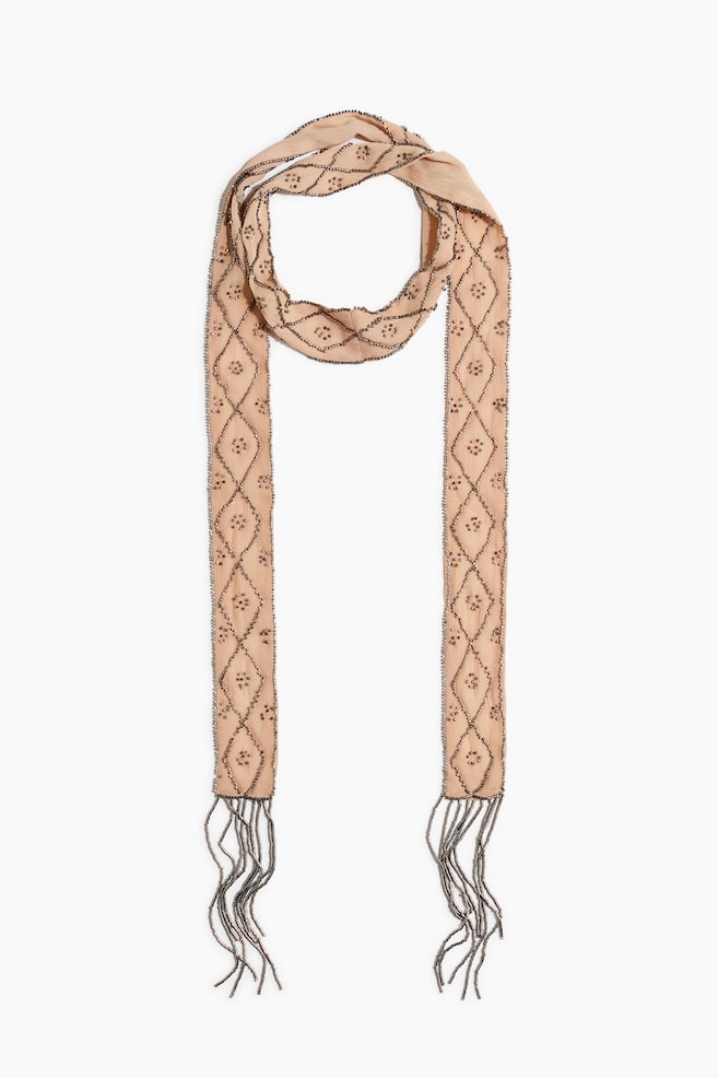 A peach-colored narrow scarf is hanging in a front view, looped at the top with two long ends draping straight down. The scarf is featuring a diamond lattice pattern embellished with dark metallic beads and small floral motifs inside each diamond, finishing with long beaded fringe at the ends. - 1