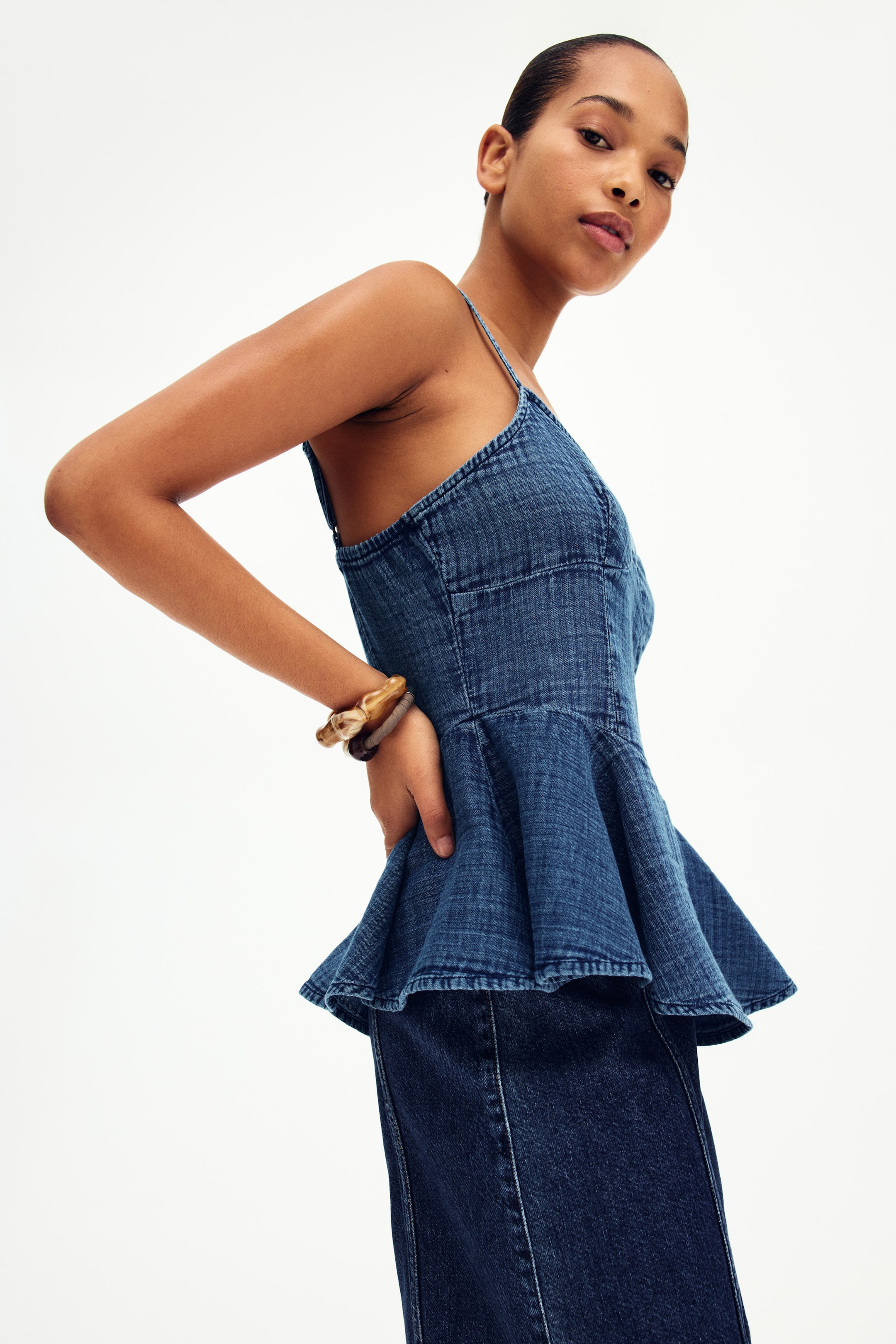 View larger image: The denim top highlights thin spaghetti straps and a snug bodice with prominent seams. It gracefully flares into a peplum hem, exhibiting a medium blue wash and layers elegantly over a darker denim bottom.