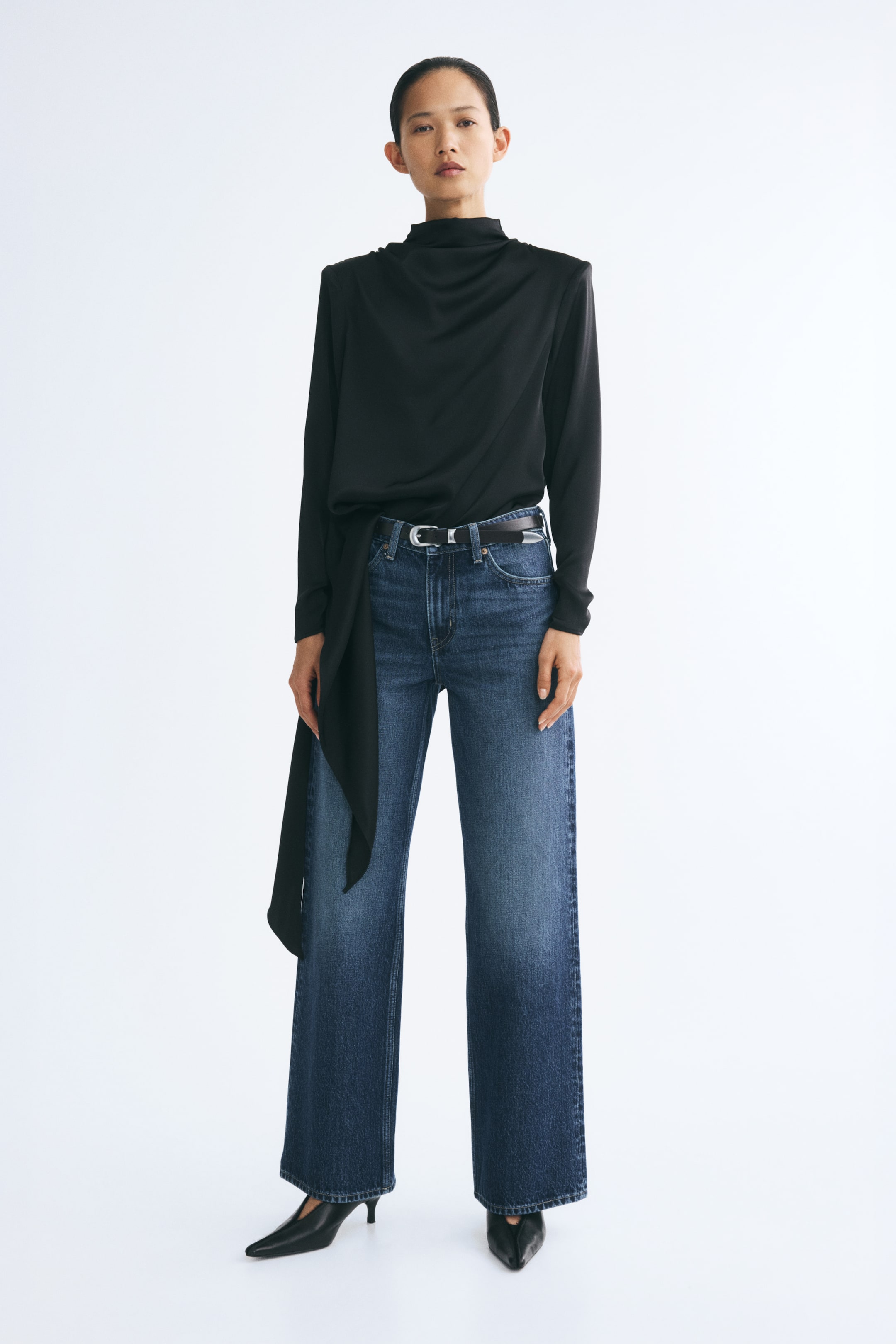 View larger image: Blue denim jeans with a wide-leg silhouette and a faded mid-wash are being worn by the model. The jeans feature a regular waist styled with a thin black belt and are paired with a draped black long-sleeved top and black pointed-toe heels.