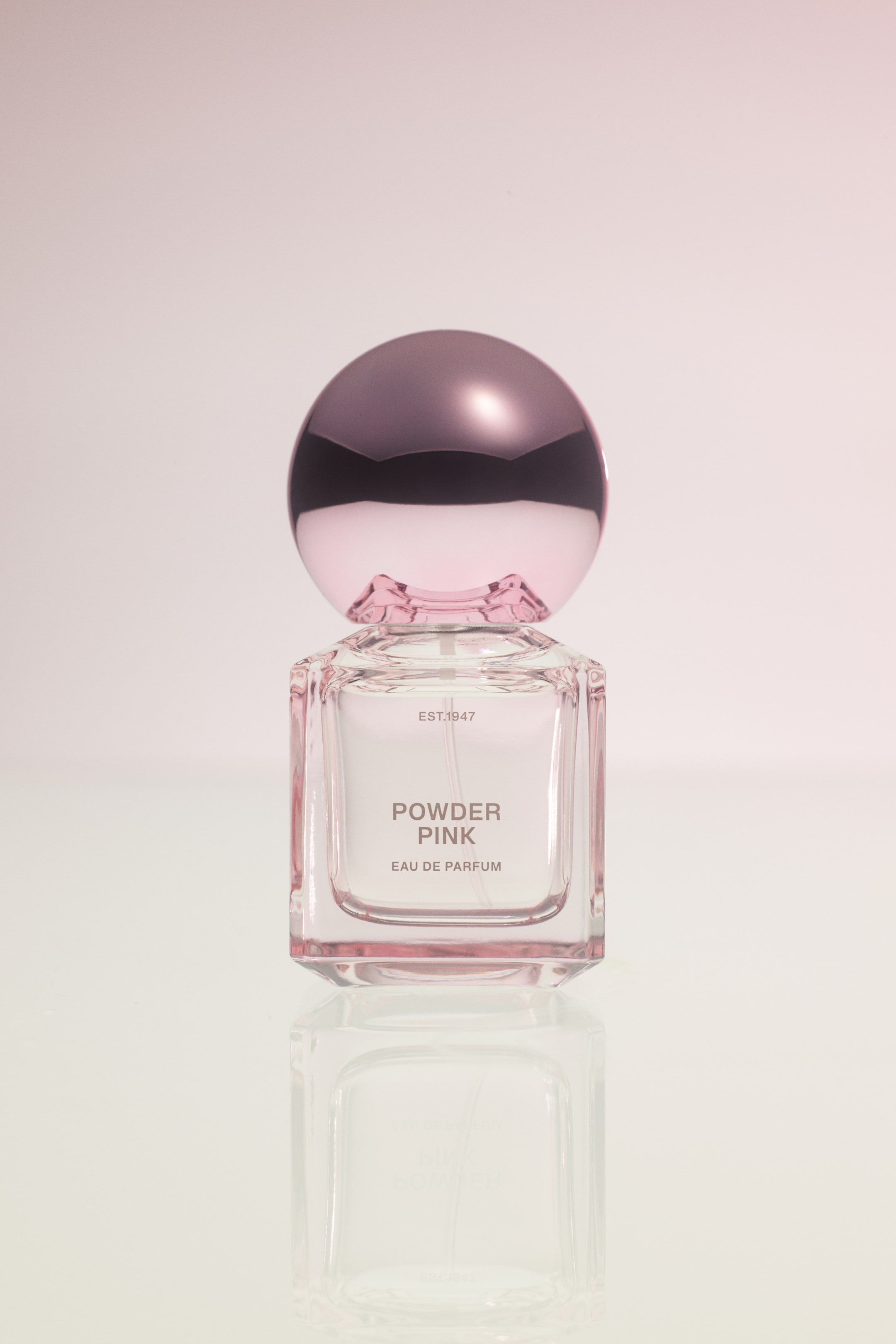 View larger image: A bottle of Powder Pink Eau De Parfum is standing upright, showcasing its square clear glass body with rounded edges and a large, metallic pink spherical cap. The front of the transparent bottle features the printed text "EST.1947", "POWDER PINK", and "EAU DE PARFUM" in a simple, grey sans-serif font.