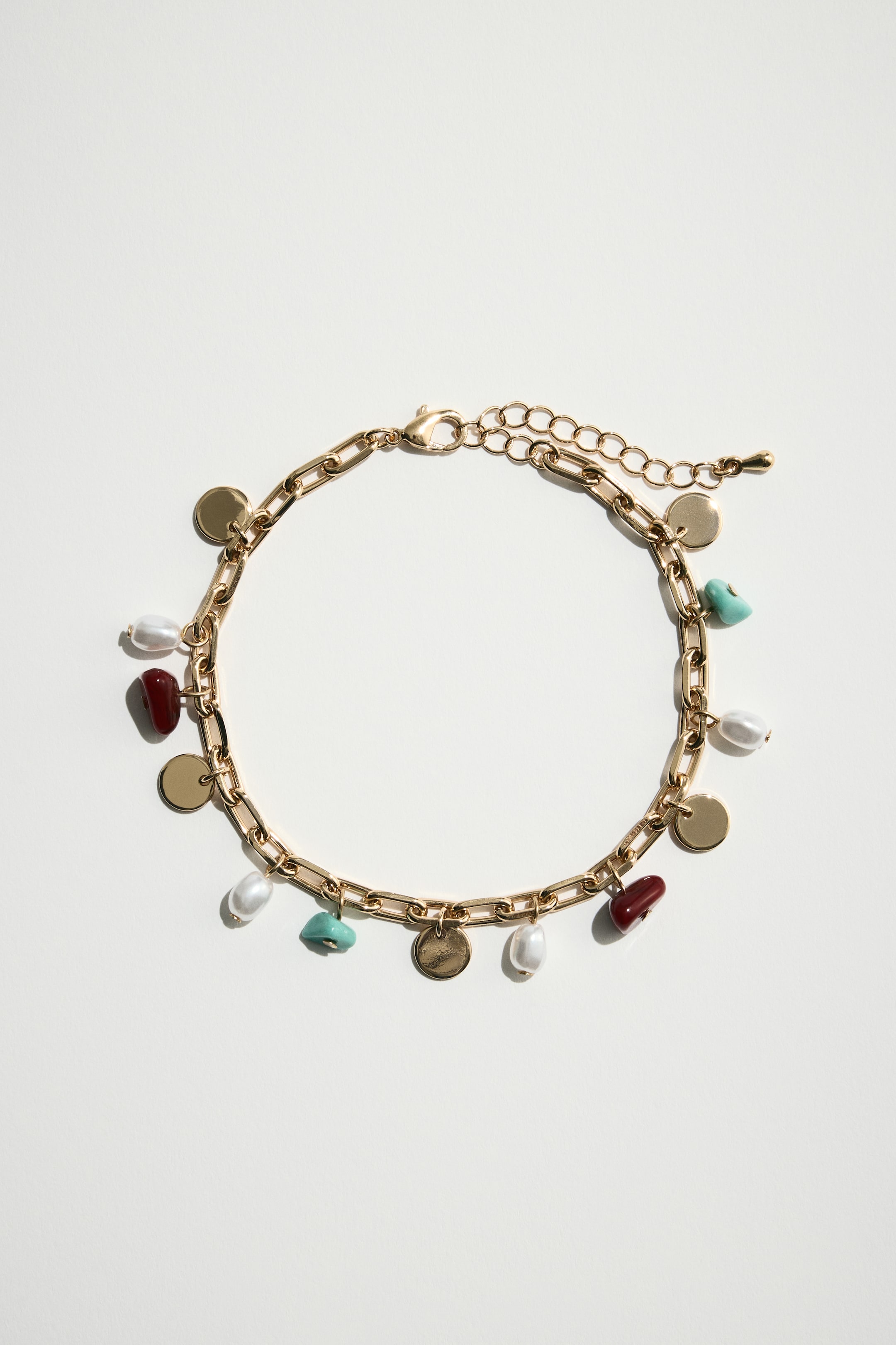 View larger image: A gold-toned paperclip chain bracelet is lying flat, forming a circular shape. It is adorned with multiple charms, including small round gold discs, irregularly shaped white pearls, dark red stones, and light blue stones. A lobster clasp and an extender chain are visible at one end of the bracelet.