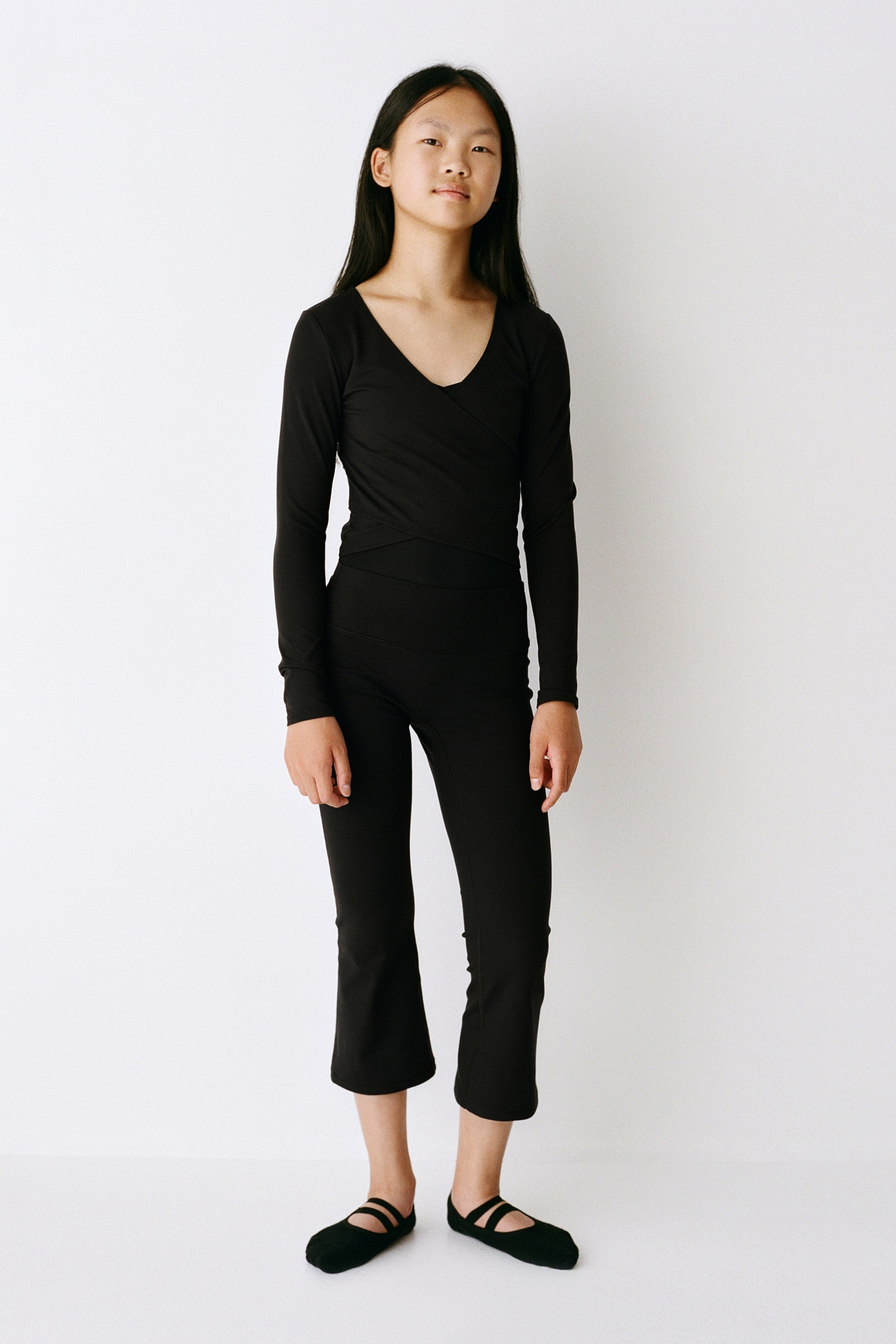 Visualizza l'immagine ingrandita: Un completo activewear nero a due pezzi è mostrato frontalmente, composto da un top avvolgente a maniche lunghe con scollo a V e pantaloni cropped svasati a vita alta abbinati. L’outfit aderente è abbinato a ballerine nere con doppio cinturino.