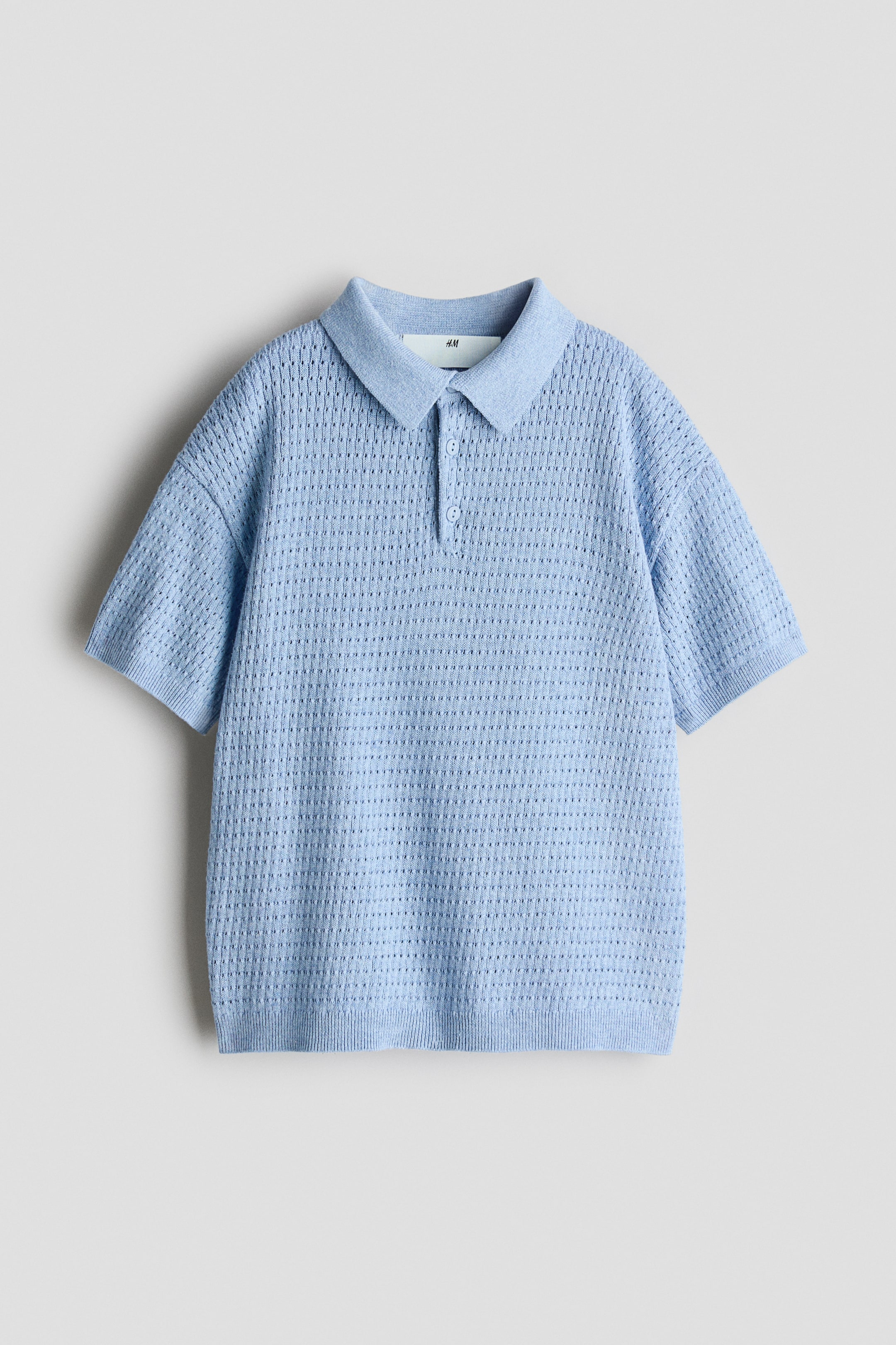 Agrandir l'image: Un polo en maille bleu clair est présenté de FACE, doté d'un col en pointe et d'une patte de boutonnage à deux boutons. Le polo présente des manches courtes avec des poignets côtelés, un motif en maille ajourée sur l'ensemble, et un ourlet côtelé.