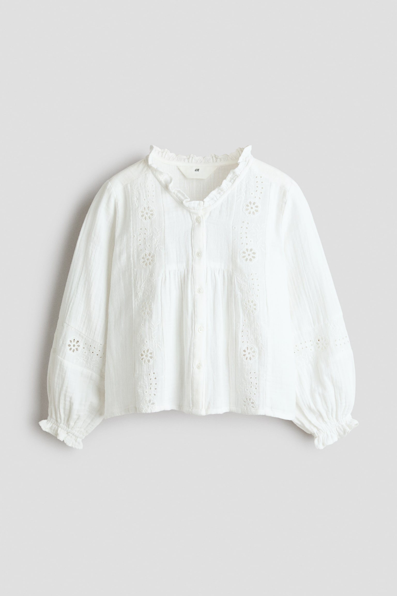 A white blouse is displaying a front view, featuring a ruffled stand-up collar and a full button-down placket with white buttons. It is showcasing broderie anglaise floral patterns on the front panels and sleeves, complemented by vertical pintuck details and elasticated ruffled cuffs. - 1