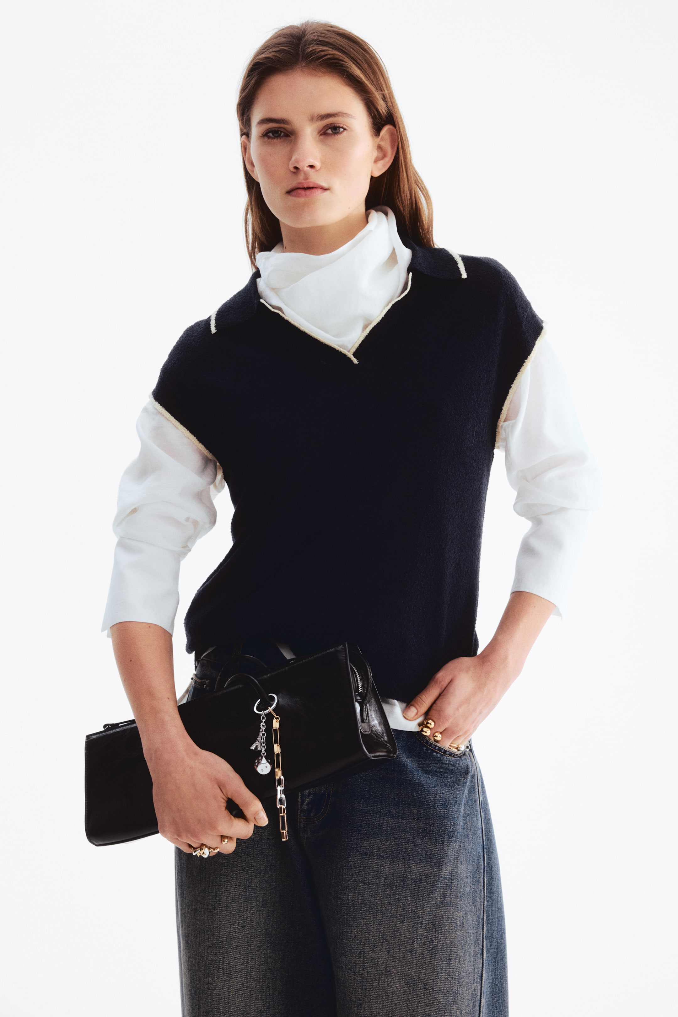 Ver imagem maior: Uma mala preta retangular e suave está a ser segurada horizontalmente e ligeiramente inclinada para a frente, mostrando um fecho de correr superior e porta-chaves decorativos com elos prateados e dourados presos a uma extremidade.