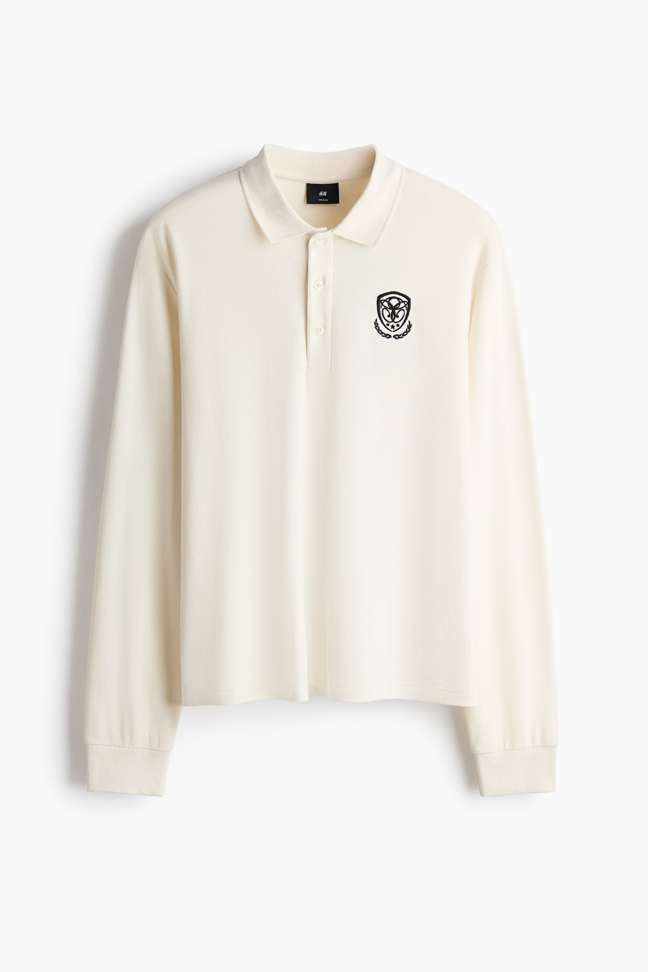 View larger image: A cream long-sleeved polo shirt is facing front, featuring a waffle knit fabric and a classic polo collar. It is displaying a two-button placket, ribbed cuffs, and a black embroidered crest on the left chest.