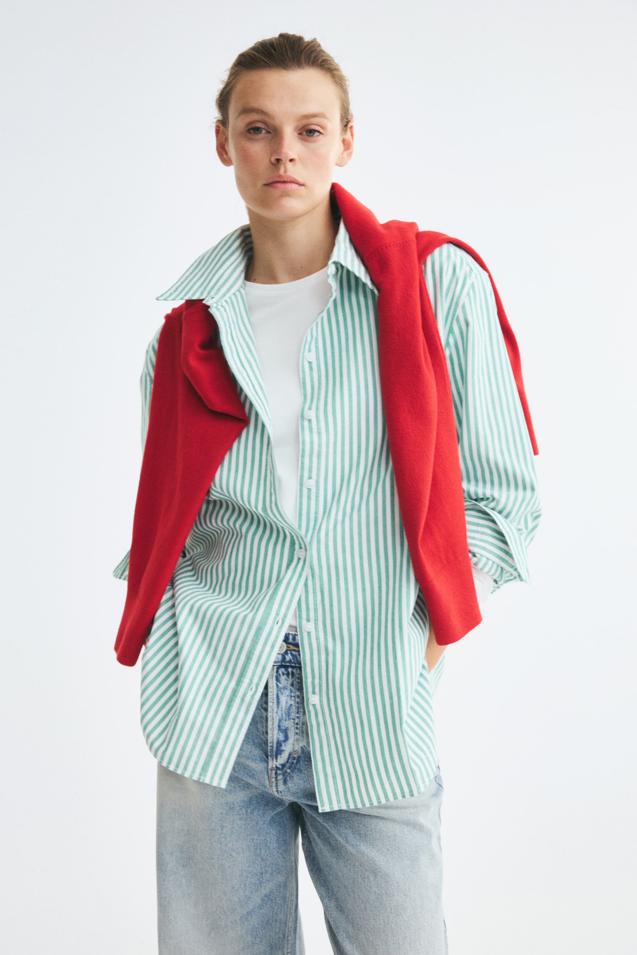 Xem ảnh lớn hơn: Một chiếc áo sơ mi dáng rộng thoải mái bằng vải cotton Oxford kẻ sọc xanh lá và trắng đang được người mẫu mặc bên ngoài áo thun trắng và quần jeans wash sáng. Áo có cổ bẻ, vai xuôi và hàng cúc phía trước, với một chiếc áo len đỏ khoác hờ trên vai.