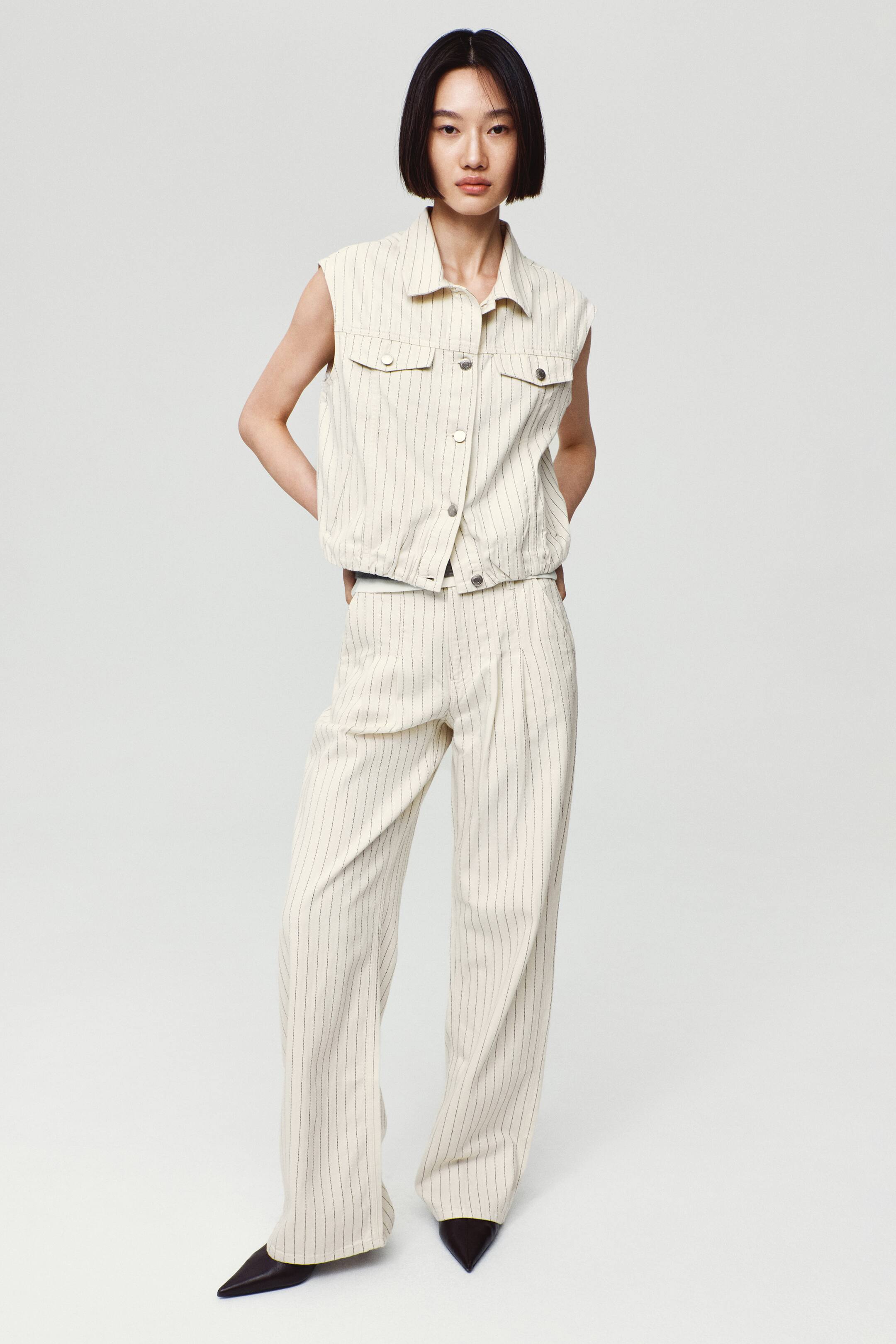 View larger image: Wide-leg, pinstriped denim jeans in an off-white shade are worn by the model, featuring front pleats and a relaxed, straight fit. The jeans are paired with a matching sleeveless pinstripe vest and black pointed-toe shoes.