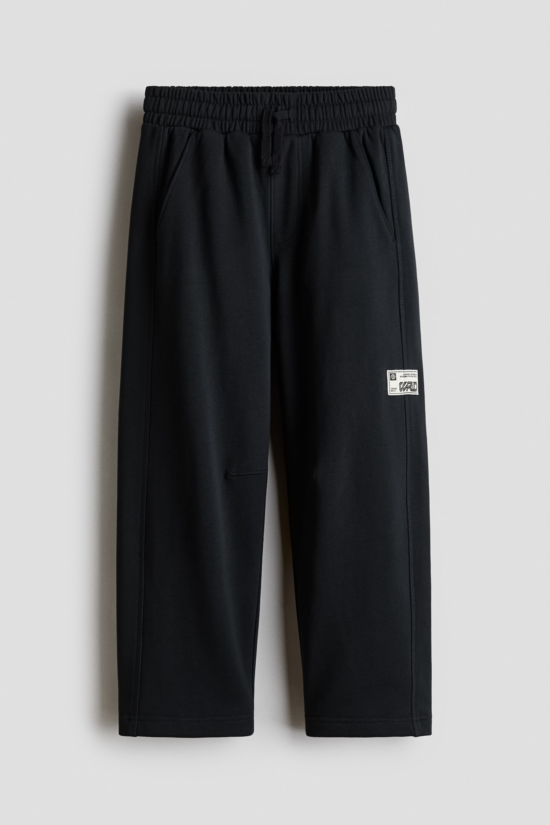 View larger image: Black sweatpants are hanging facing front, featuring an elasticised drawstring waistband, side slip pockets, and a relaxed, wide-leg silhouette. The pants display a small white rectangular graphic patch on the outer side of the left leg and straight, open hems.
