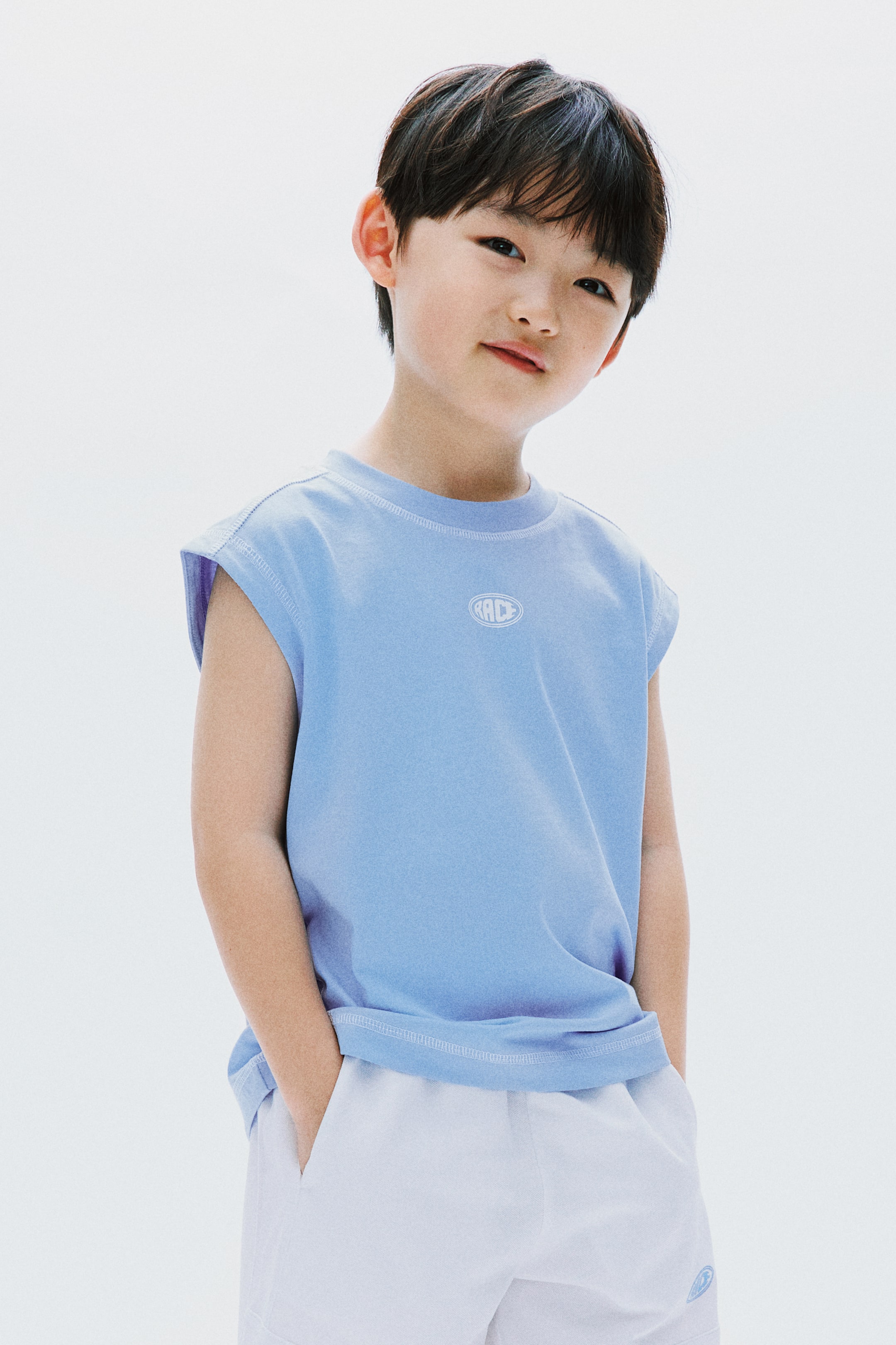 拡大画像を表示: ライトブルーのノースリーブTシャツは、クルーネック、肩、アームホール、裾にホワイトのコントラストステッチが施され、左胸にはホワイトの楕円形の『RACE』ロゴが付いています。ホワイトのショーツと組み合わせており、右脚下部にライトブルーのエンブレムが表示されています。