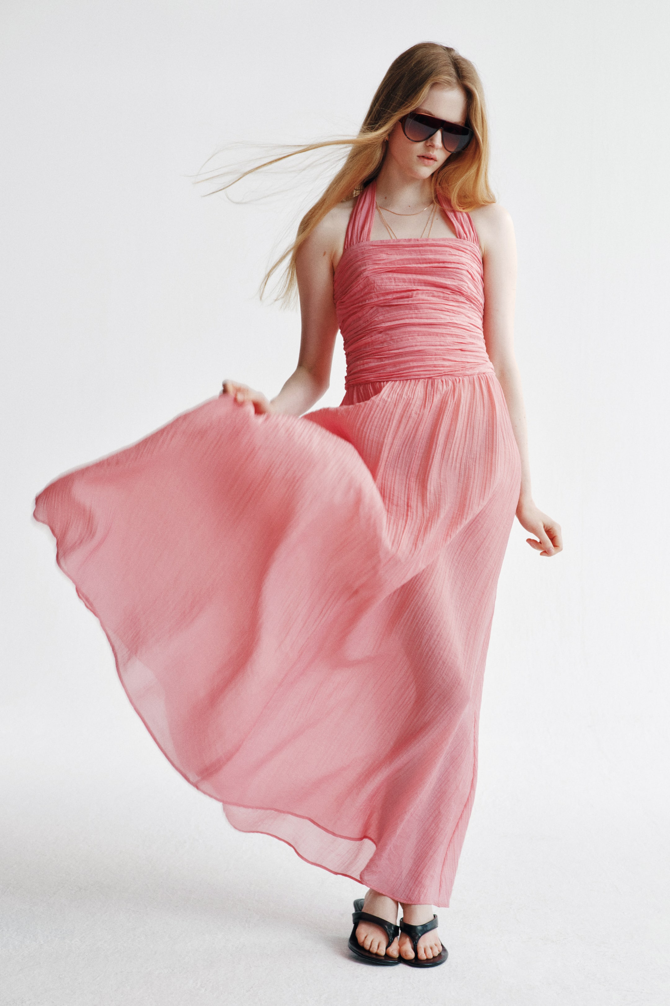 Grotere afbeelding bekijken: Een lange roze jurk van een doorschijnende, gekreukte stof wordt gedragen door het model. De jurk heeft een aansluitend, geplooid lijfje met een rechte halslijn en brede halterbanden, een geplooide taille naad en een volumineuze, uitlopende rok. Het model houdt de rok vast, waardoor deze opbolt.