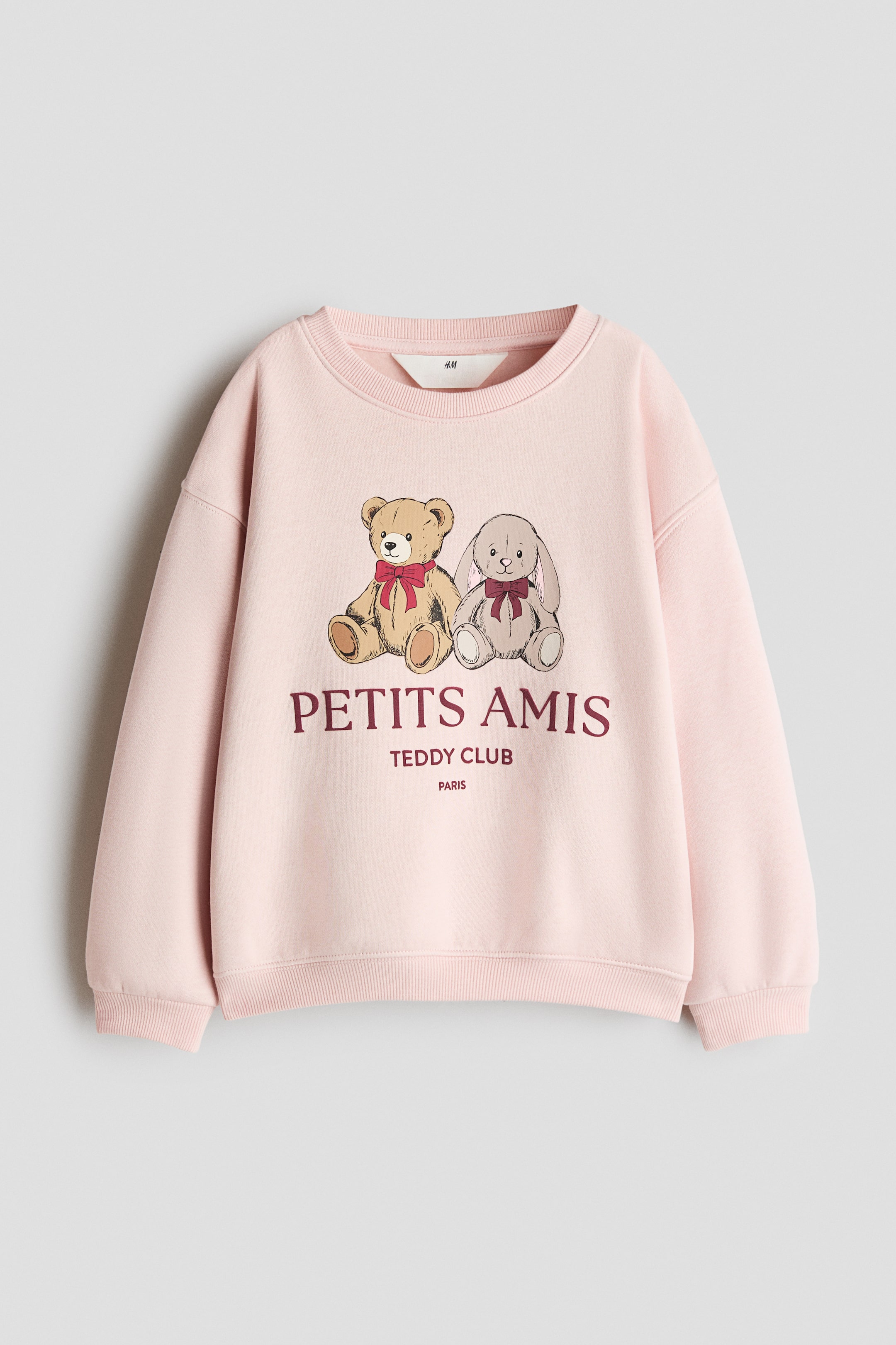 View larger image: A light pink sweatshirt is shown from the front, featuring a round ribbed neckline, dropped shoulders, and ribbed cuffs and hem. The front displays a printed graphic of a teddy bear and a rabbit wearing bows, with the burgundy text "PETITS AMIS TEDDY CLUB PARIS" printed underneath.