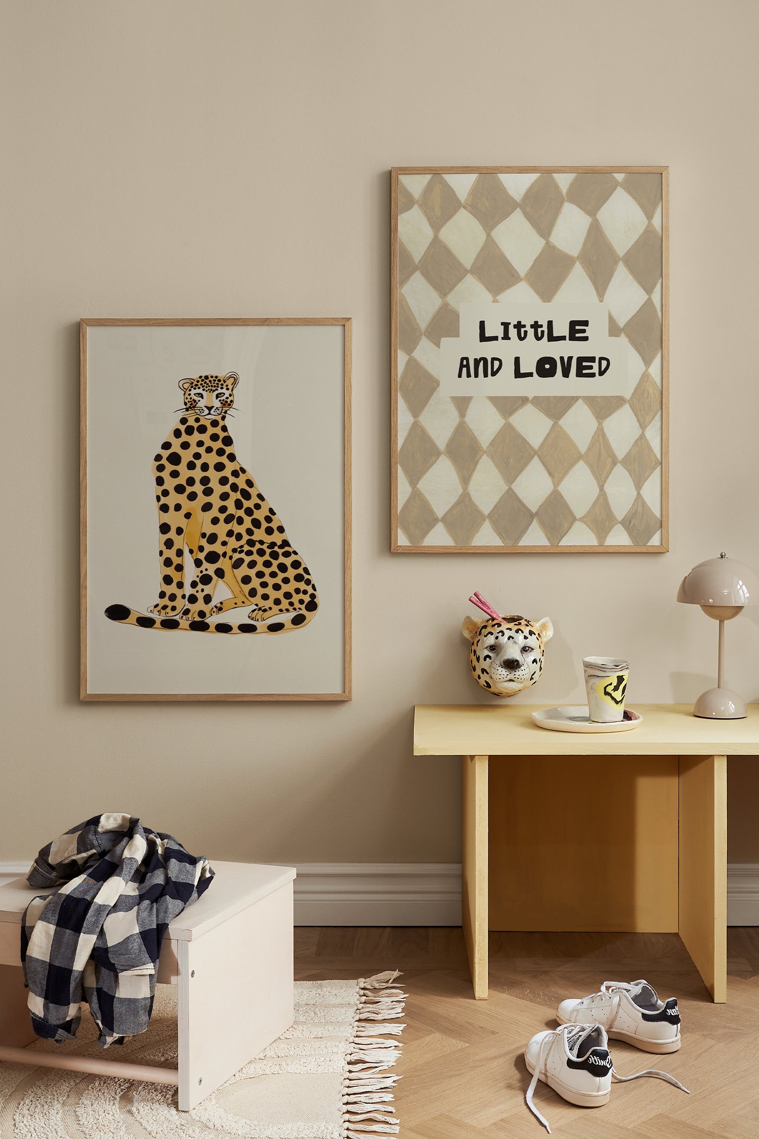 Uma impressão artÃstica retangular apresenta um padrão argyle pintado à mão em tons suaves de bege e creme. Um bloco em tom creme no centro exibe o texto "LITTLE AND LOVED" numa fonte preta, em negrito, desenhada à mão.