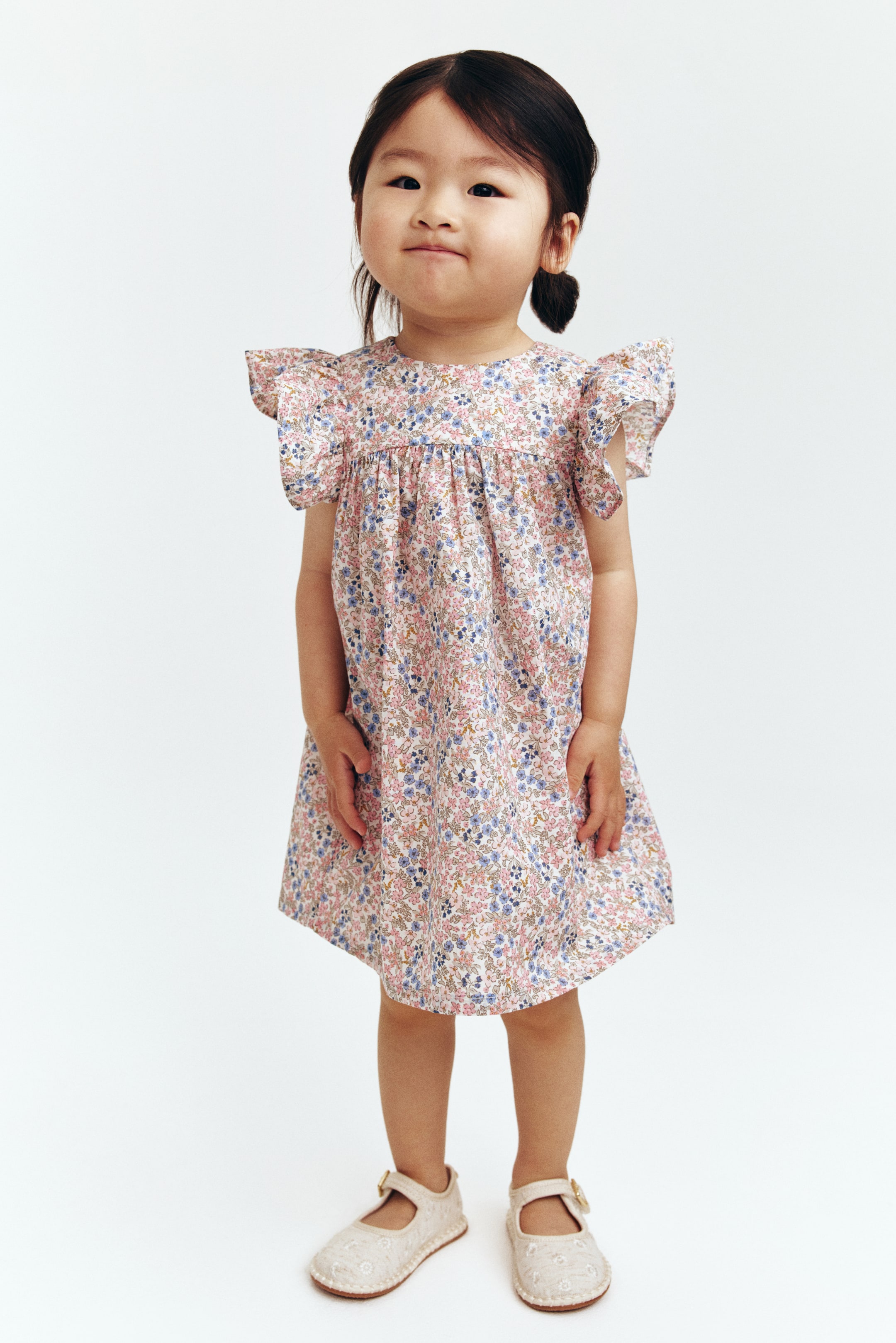 View larger image: A child's frock is presenting a soft pink base adorned with a comprehensive floral print in hues of pink, blue, and green. It comes with a round neckline, short ruffled sleeves, and a gathered skirt that flares from the waist seam.
